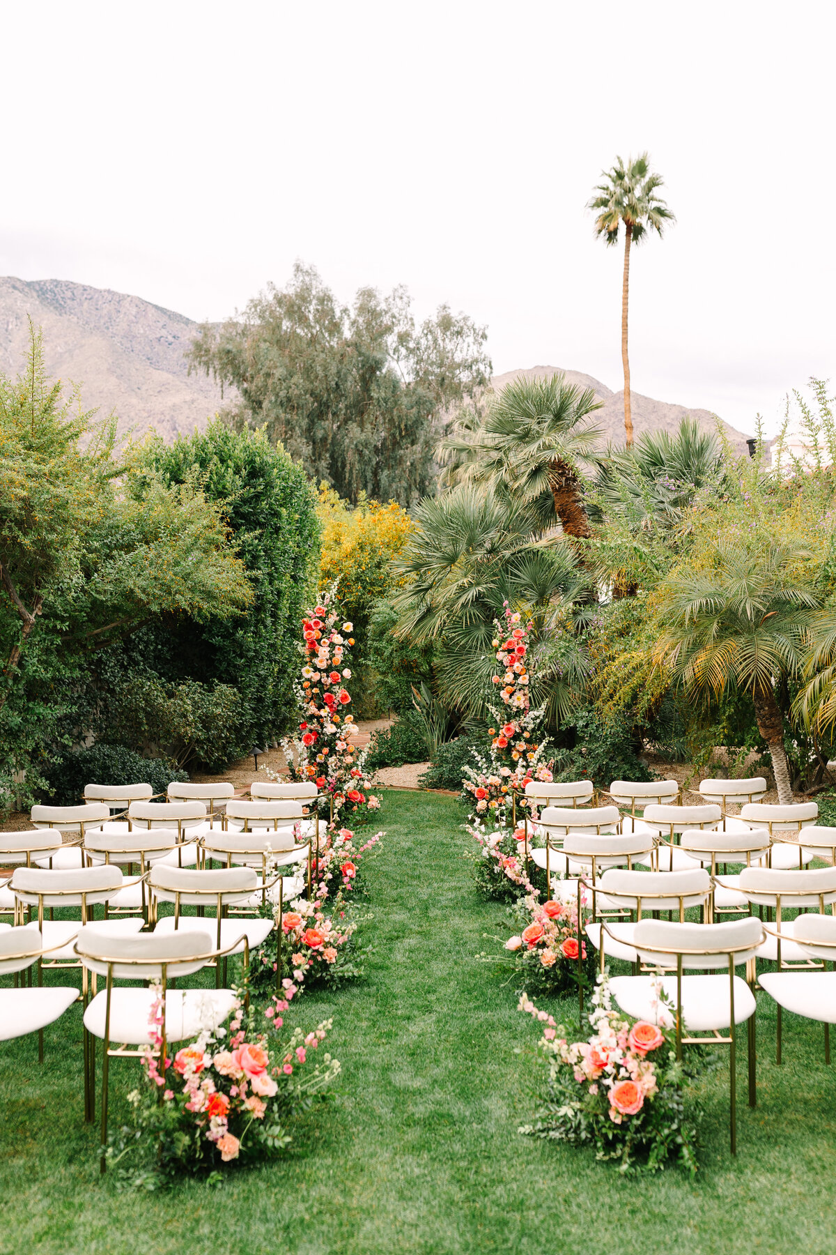 sonoma wedding ceremony set-up with white mid-century modern chairs and pink and orange florals with palm trees in background.