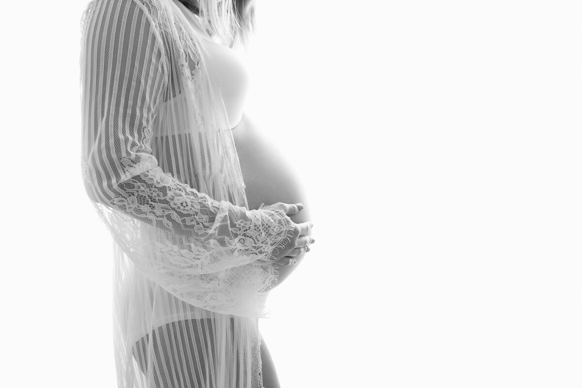 High key maternity portrait of a woman wearing a white lace gown with her hands on her belly.