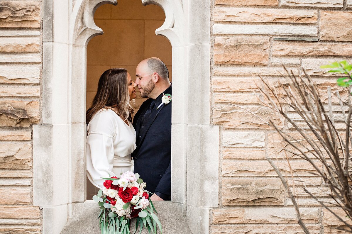 The bride, wearing a long sleeved wedding dress leans in to kiss the groom who is wearing a black suit with a white boutonniere. The bride is holding a blush and red bouquet with greenery as they are framed in a gothic window surrounded by a light brown sandstone building at Scarritt Bennett.