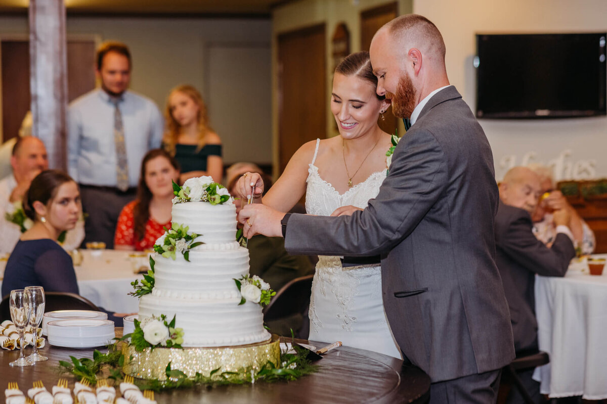 photo of a bride and groom cutting and white cake with white flowers on it as people look in the background