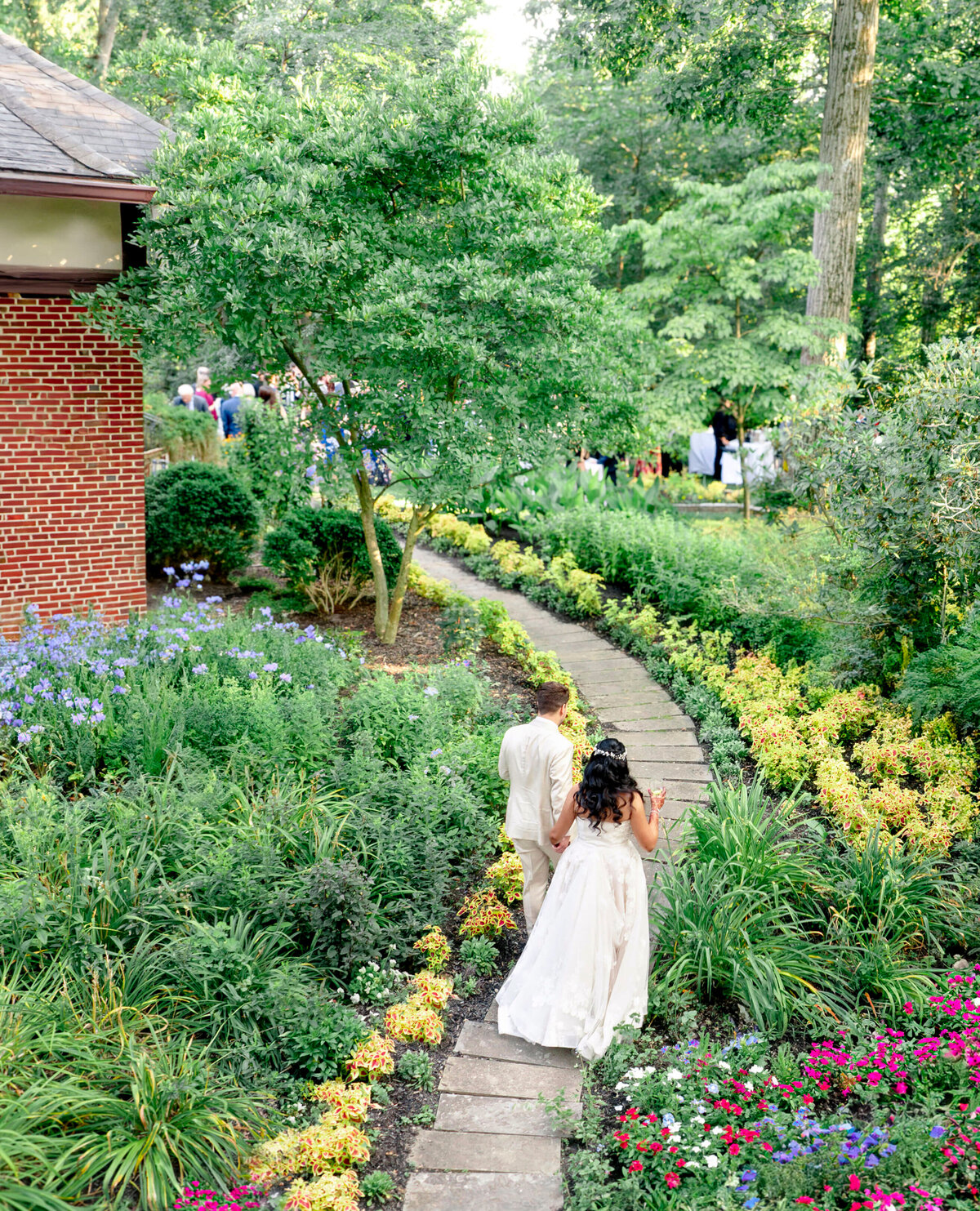 A bride and groom walk hand in hand down a stone path surrounded by lush green gardens. The bride wears a white dress, and the groom is in a light suit. Guests are gathered in the background near a brick building.