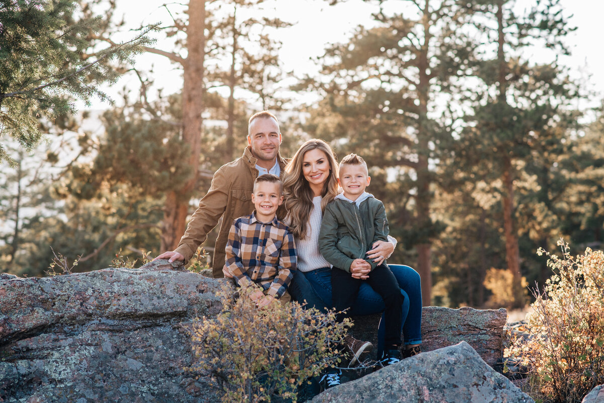 denver family photographers captures young family sitting on a boulder int he rockies while the sun shines behind them through the tall pine trees