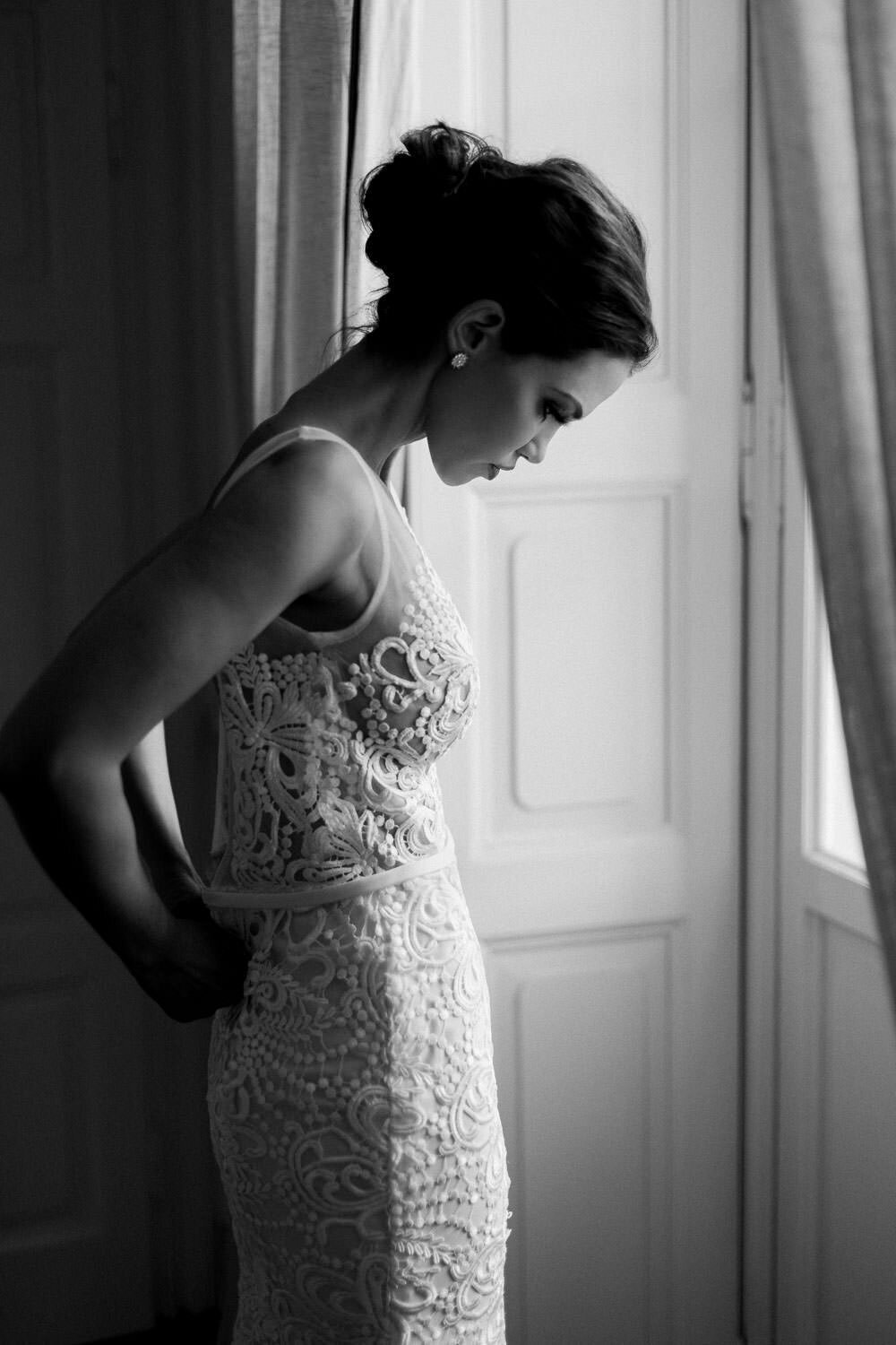 bride bride putting on white lace wedding dress by window before wedding ceremony in Lake Como Italy on white lace wedding dress by window before wedding ceremony in Lake Como Italy