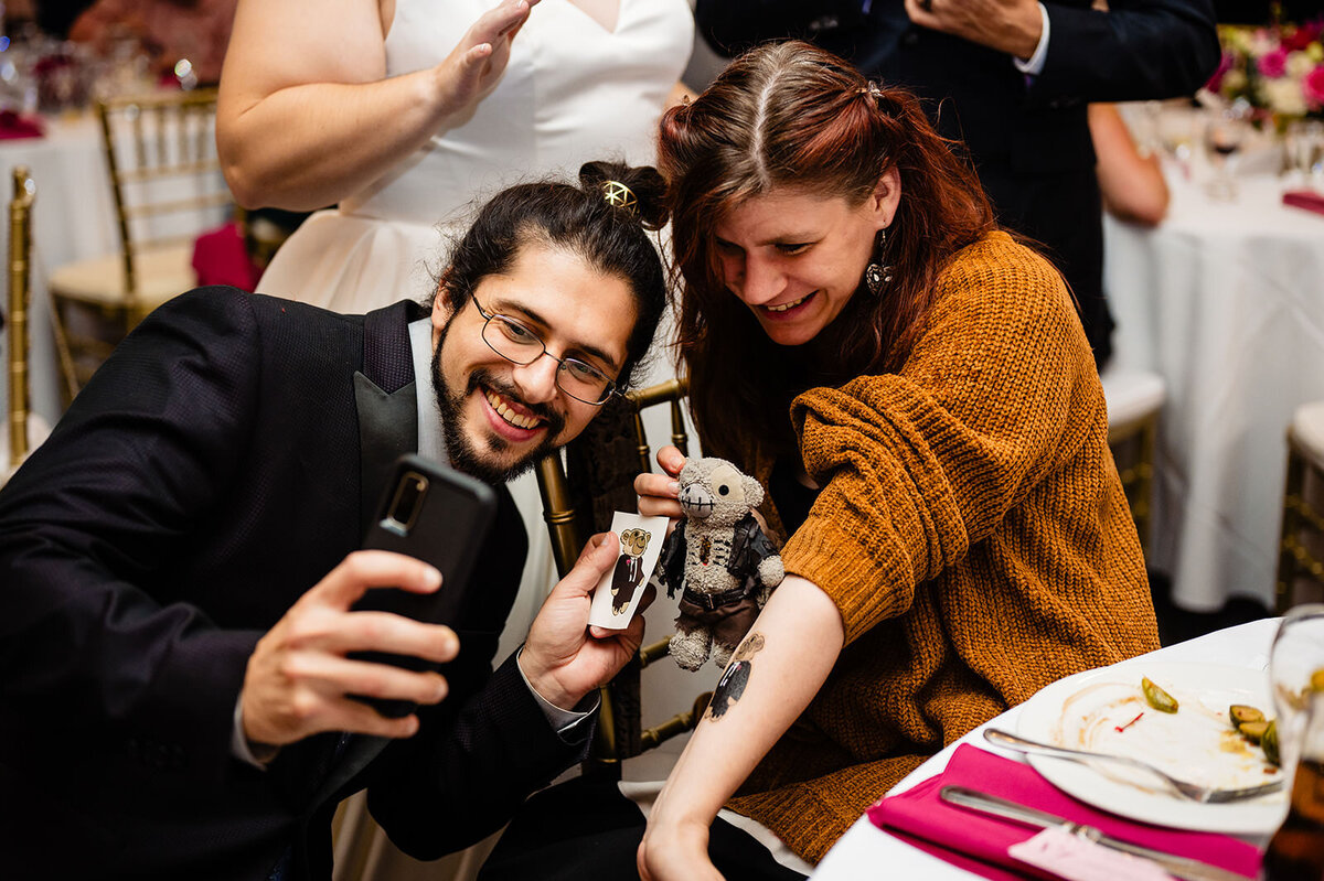 A couple takes a selfie with a small, crocheted toy, celebrating at a wedding reception with guests in the background