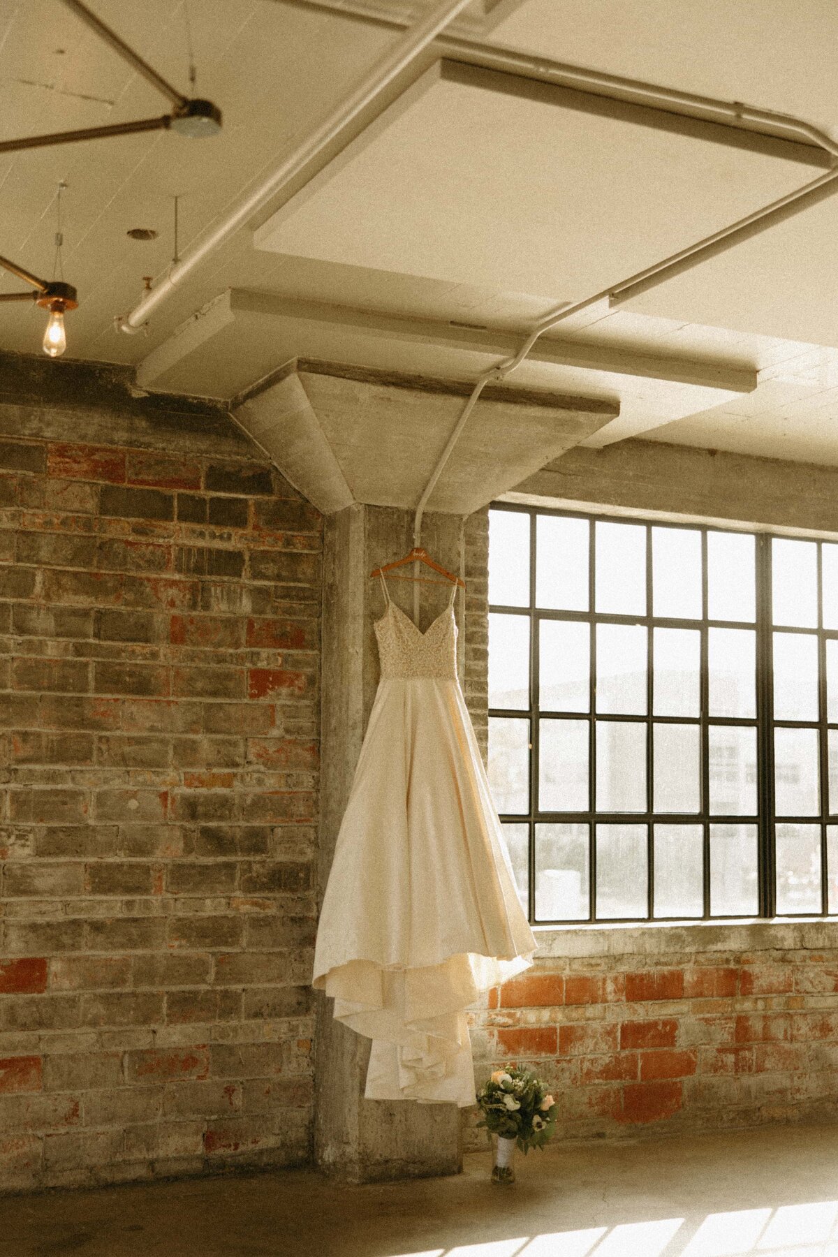 A wedding dress hangs from a beam in a loft with brick walls and a large window, bathed in natural light, perfect for Iowa weddings.