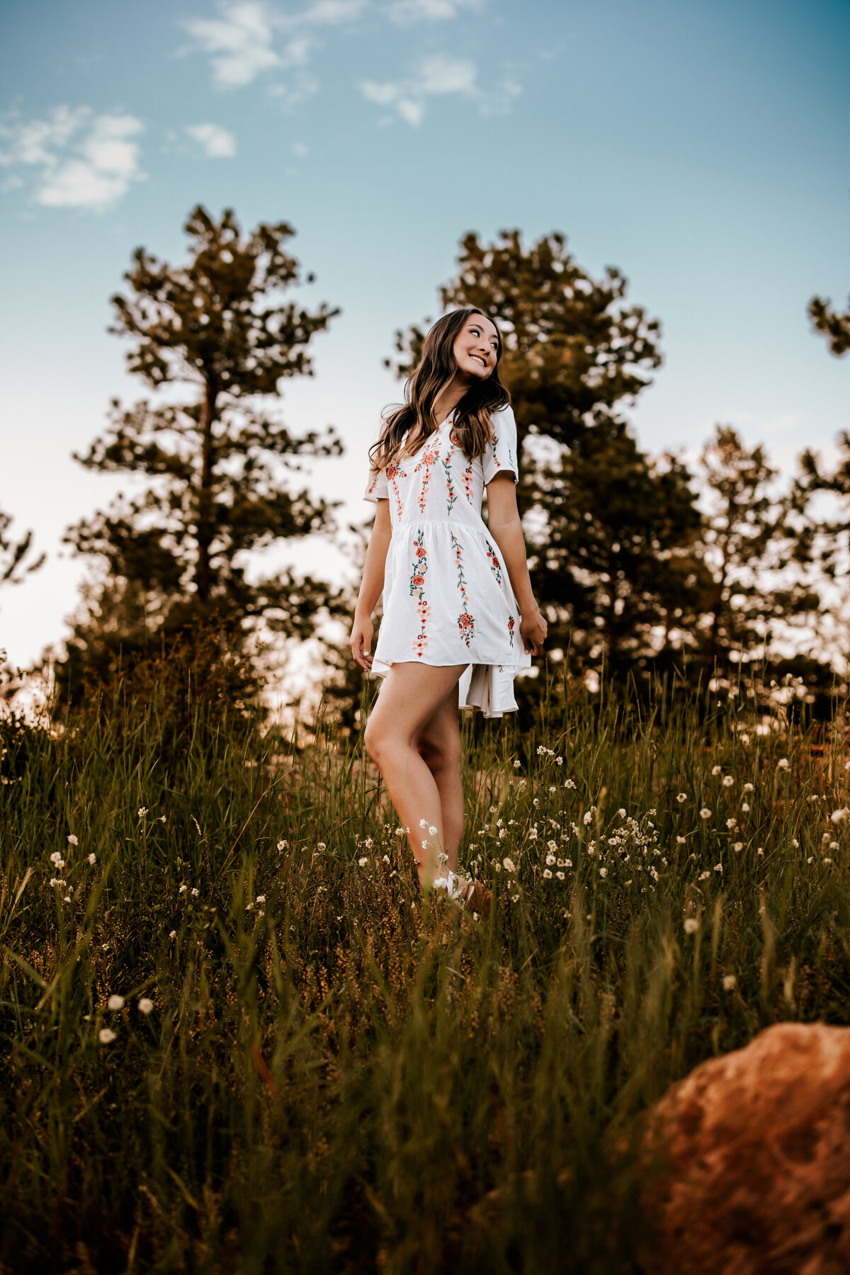 girl poses in a field of small blooming flowers and pine trees for senior photos