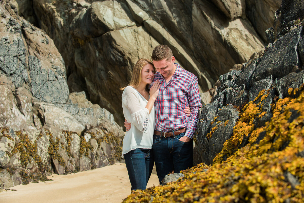 young couple embracing during engagement portrait while standing on the beach surrounded by rock