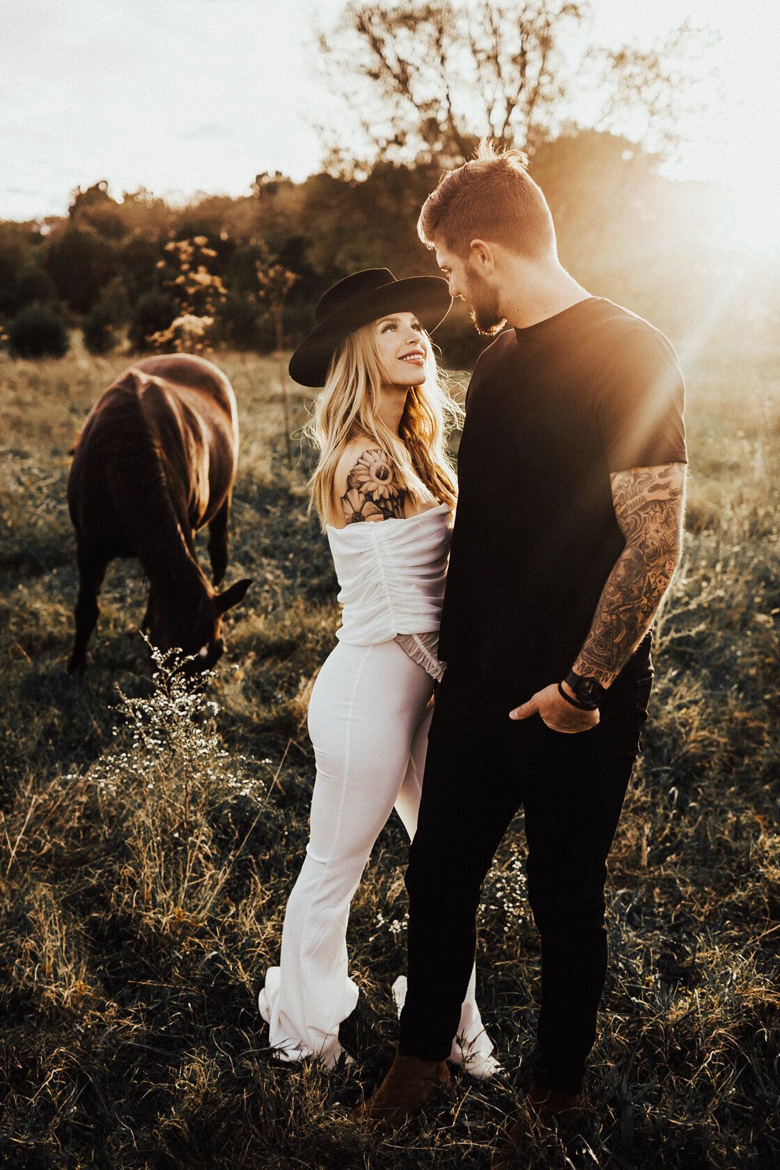couple holding hands and smiling at each other with horses in a field