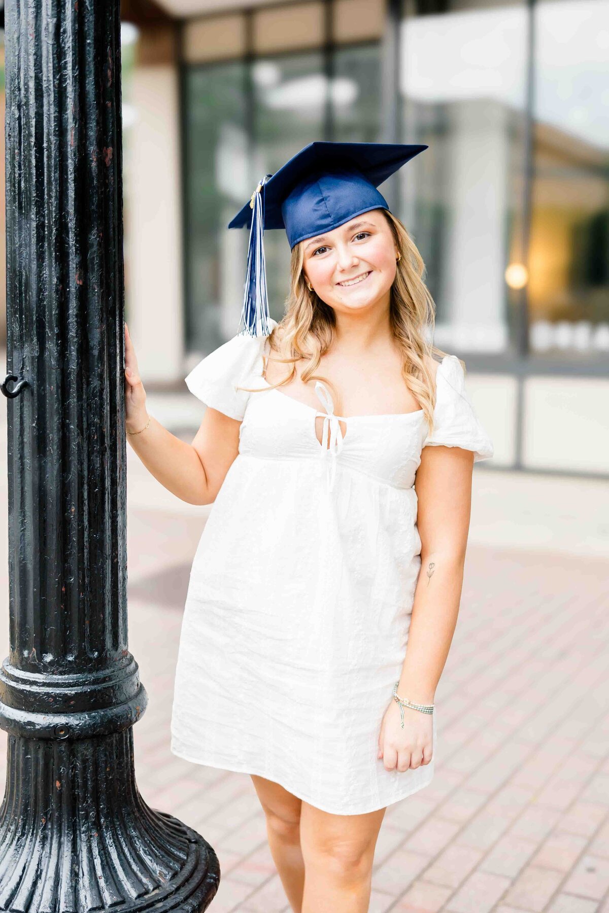 A person wearing a white dress and blue graduation cap stands smiling beside a black lamppost, captured perfectly by a senior photographer in Fayetteville, NC. They are outdoors, in front of a building with large windows.