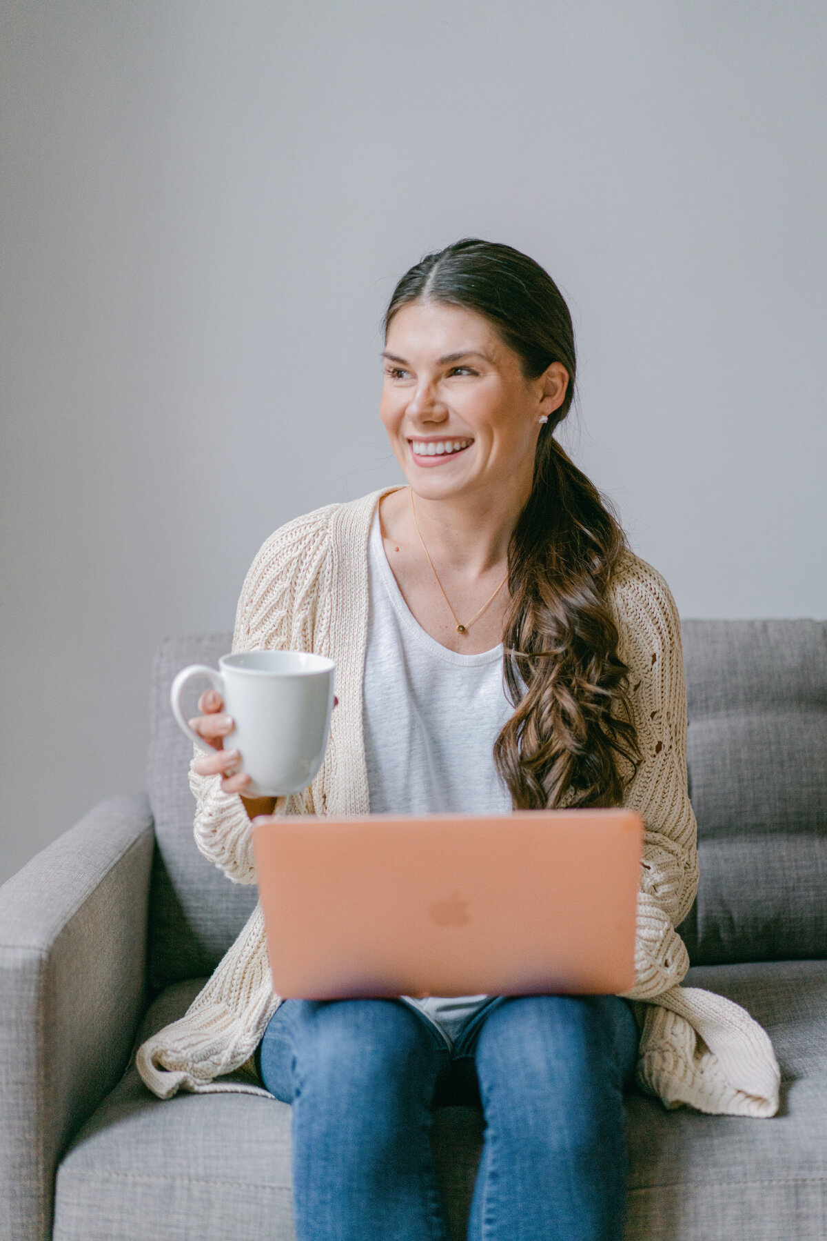 woman with a coffee cup in one hand and a laptop on her lap looking away from the camera and laughing