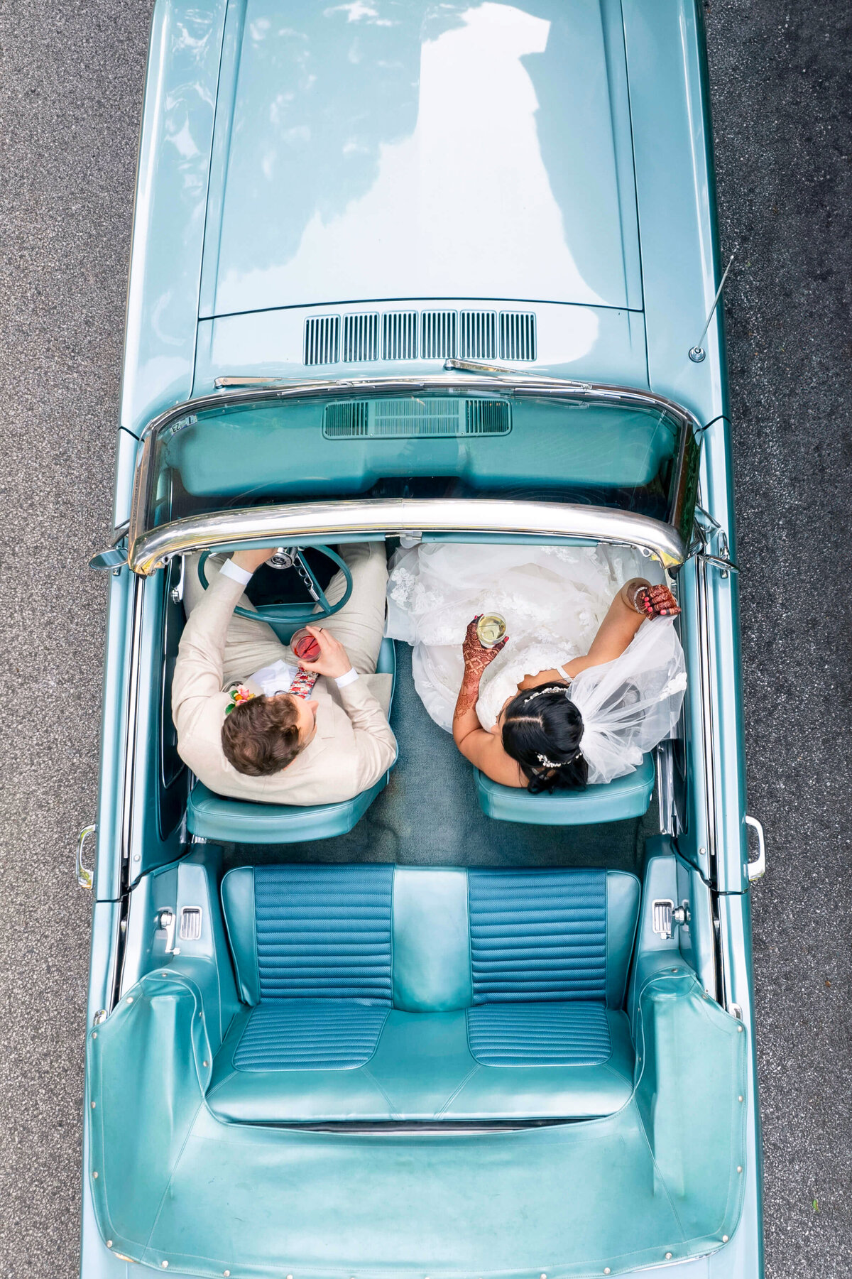 Aerial view of a couple in wedding attire sitting in a blue vintage car. The groom is in a light-colored suit, and the bride is in a white dress. The car is parked on a paved road.