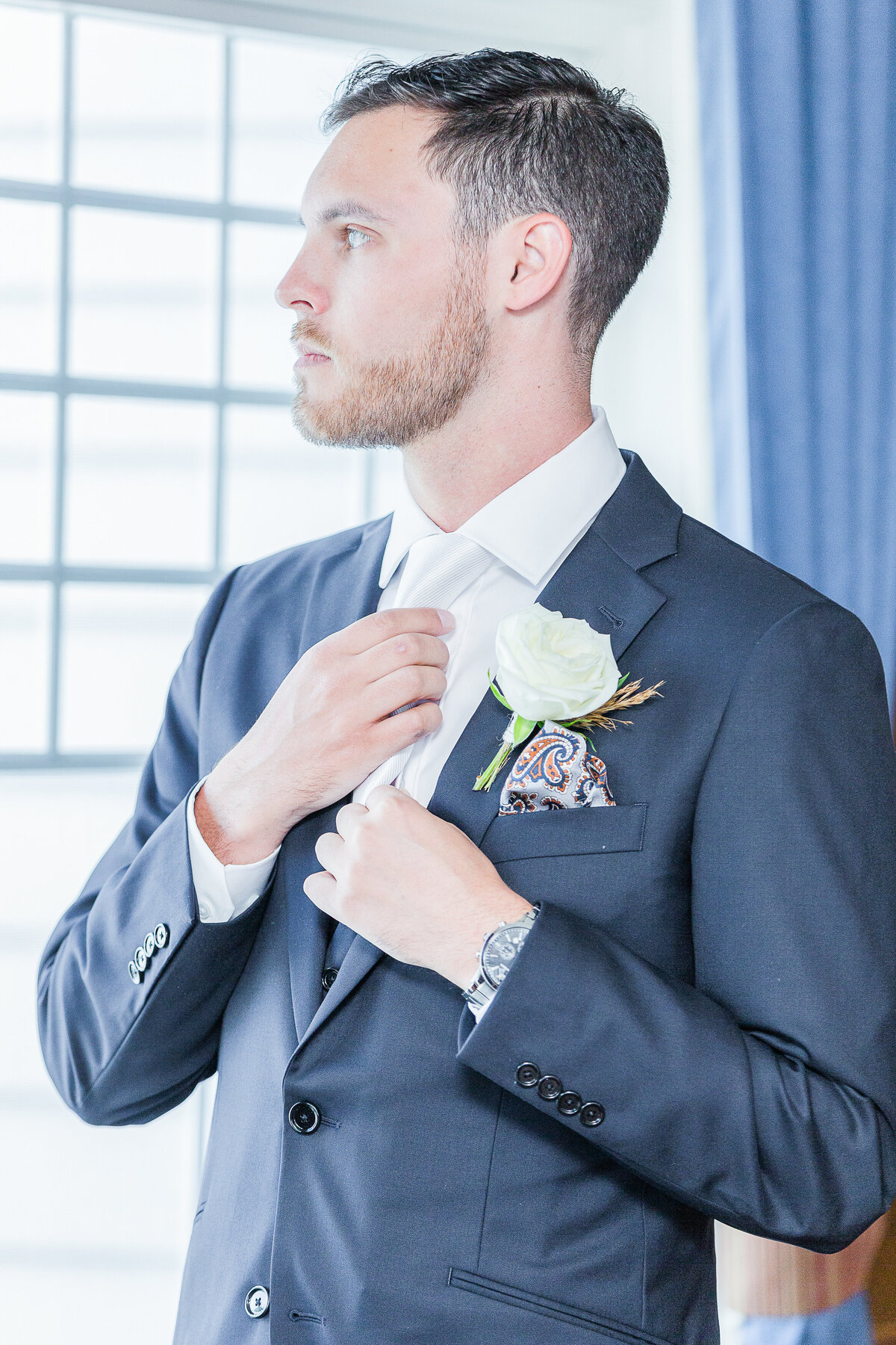 Groom adjusts his tie as he is looking out the window  with a serious expression. Captured by best New England wedding photographer Lia Rose Weddings.
