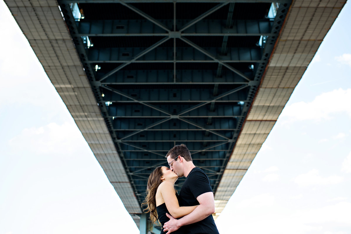 Two people in love kiss under the ben franklin bridge.
