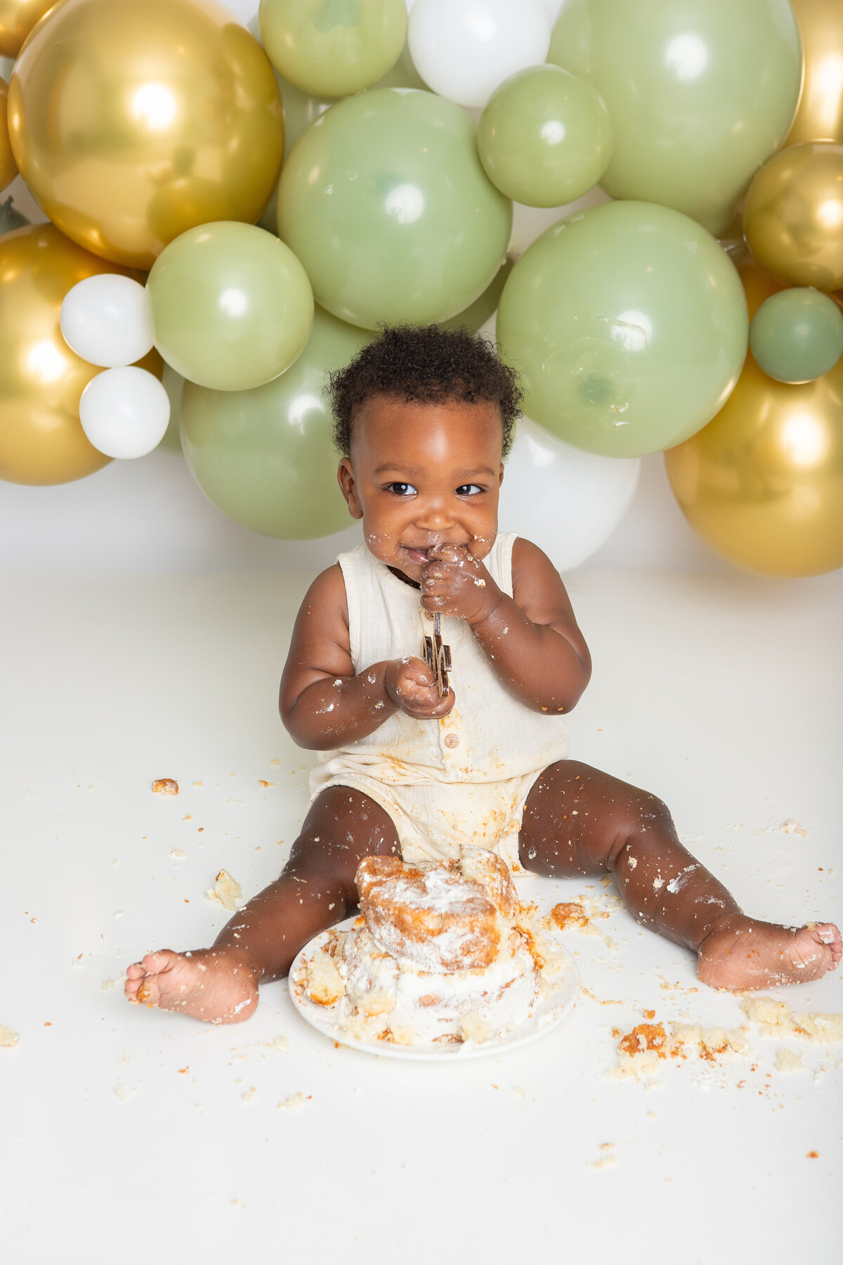 Baby eating a simple white cake with an olive and gold balloon backdrop