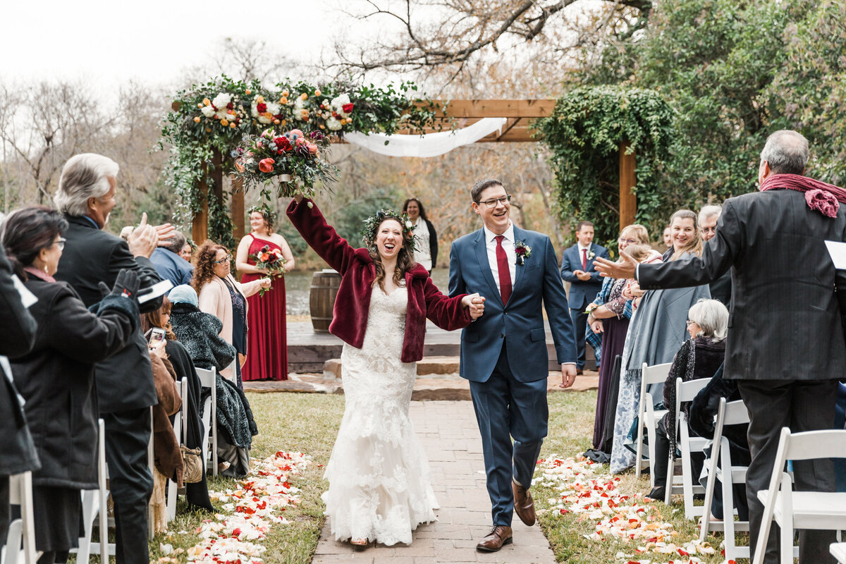 A bride and groom joyously celebrating as they recess down the aisle after their wedding ceremony at Cross Creek Ranch in Parker, Texas. The bride is on the left and is wearing an intricate, white dress, flower crown, and red jacket while holding up a large bouquet high into the air in celebration. The groom is on the right and is wearing a navy suit, red tie, and boutonniere. Guests stand, applaud, and cheer on either side of the aisle. Behind the couple stands the officiant and wedding party who are all backed by a large wooden arch frame covered in flowers and floral arrangements.