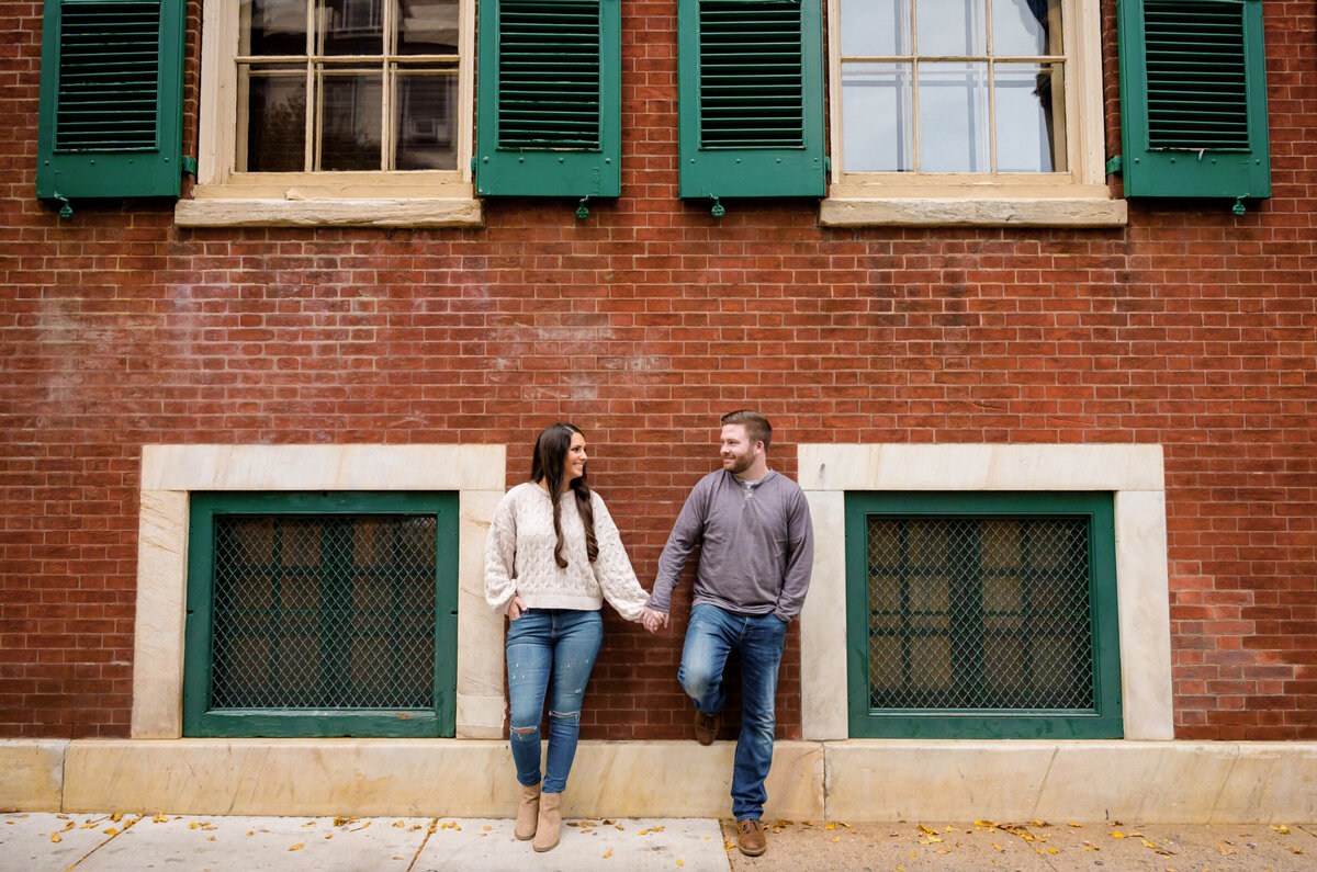 Woman with long brown hair, holding hands with a man while they lean against a brick building that has windows with green shutters and Philadelphia PA