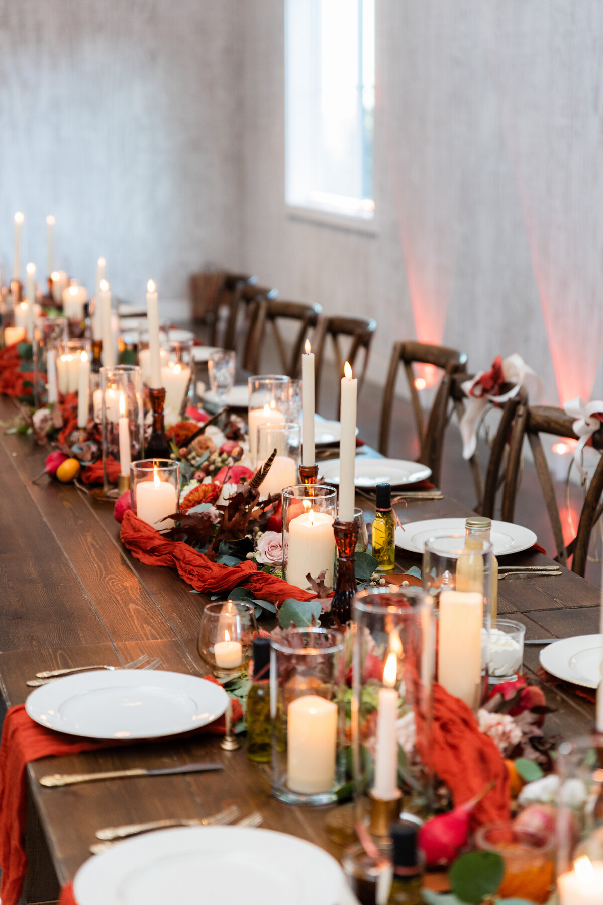 A table scape features various candles, a red tulle runner and warm colored florals.