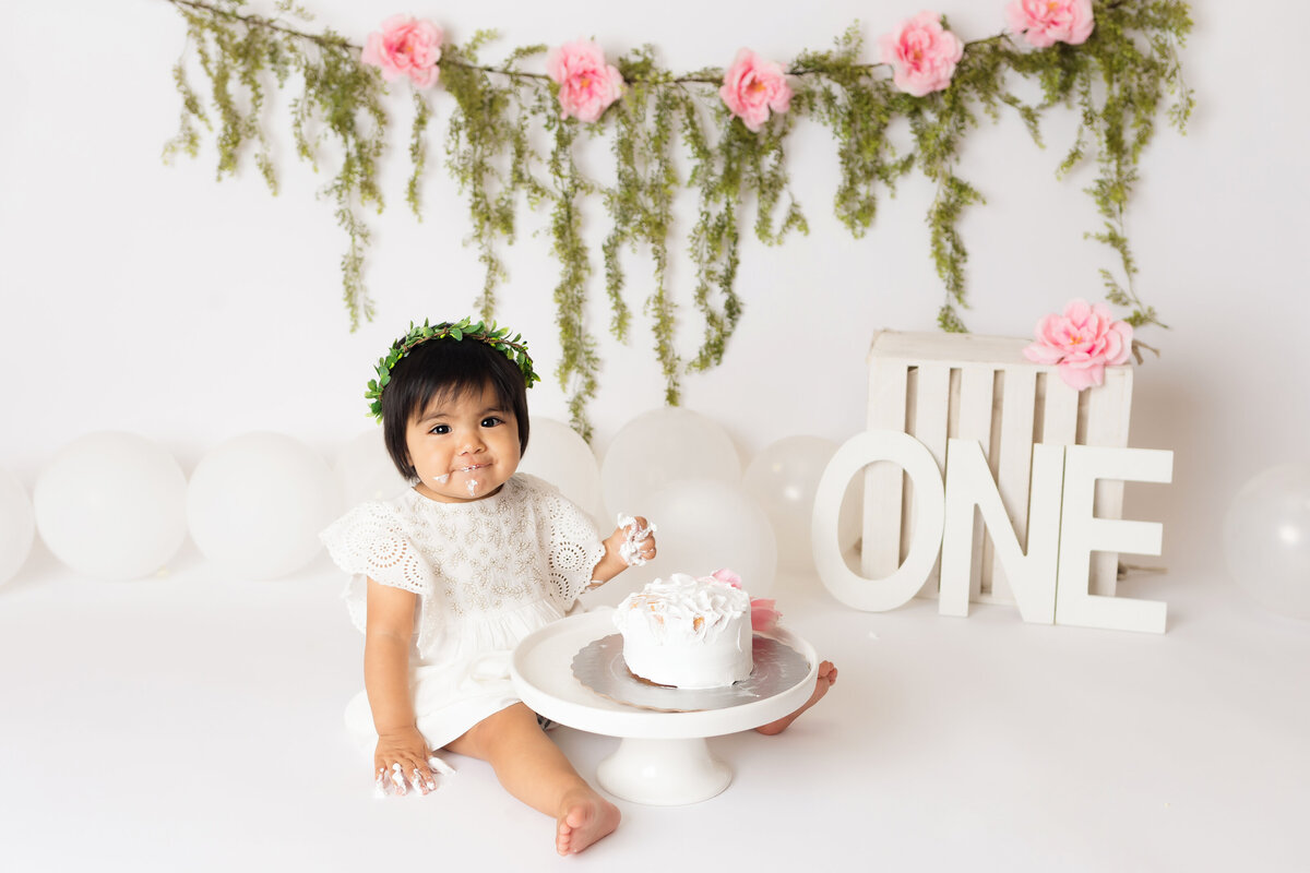 Cake Smash Photographer, a baby girl eats cake before a mossy backdrop with flowers