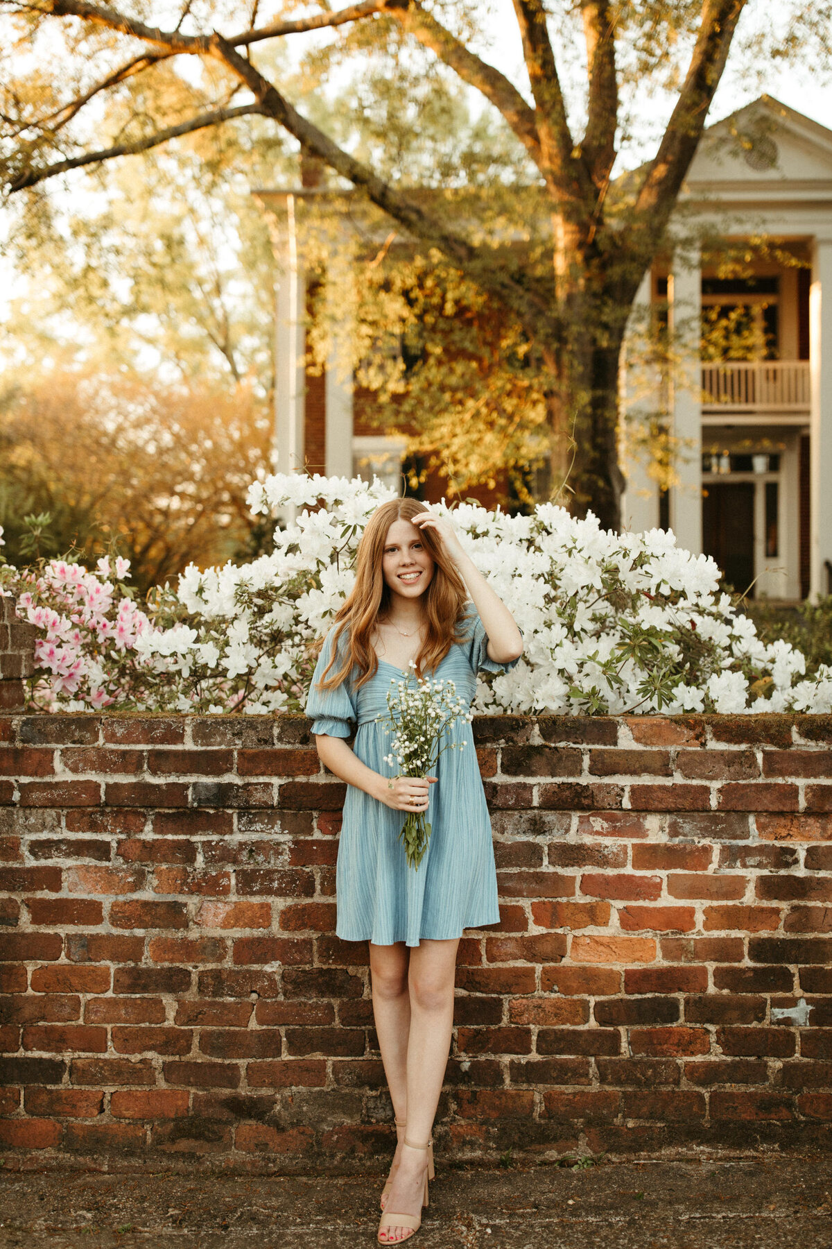 Girl senior in short blue dress standing in front of a brick fence on a sidewalk downtown