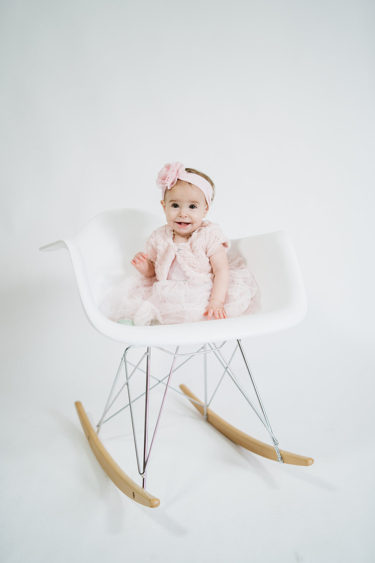 Baby toddler sitting in Eames rocking chair wearing pink dress in San Antonio photo studio on a white background