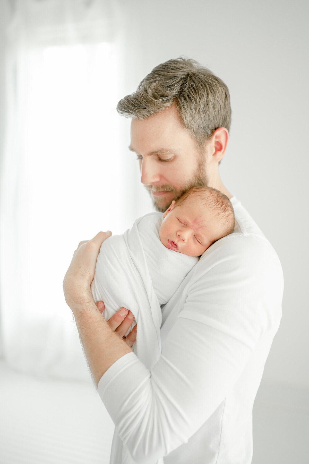 A man in a white shirt holds his sleeping baby In Kristie Lloyd’s Nashville newborn photographer studio
