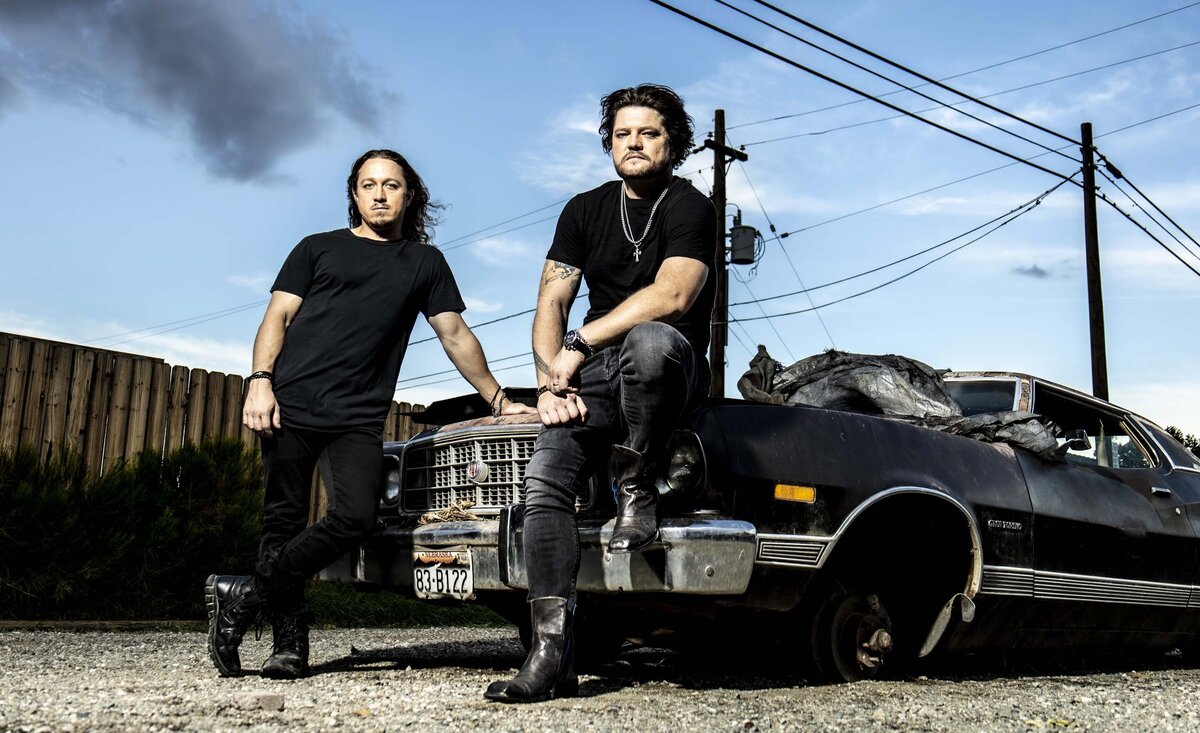 Musical duo Radiofix sitting against broken down black car power lines with smoke behind