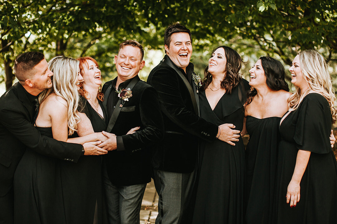 Two grooms wearing black tuxedos share smiles outside with their wedding party also wearing all black.