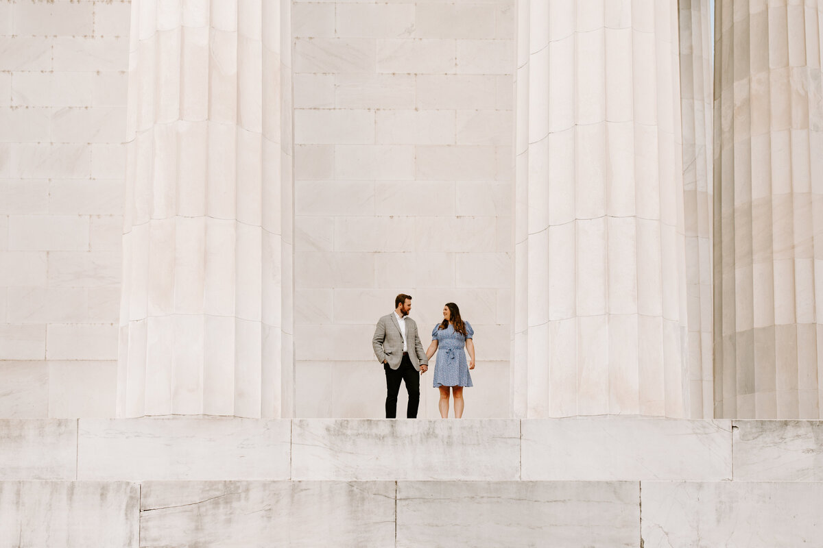 A male and female coupld stand hand in hand while standing in front of a DC monument. It is a wide shot and the couple is surrounded by beautiful white stone architecture and pillars.