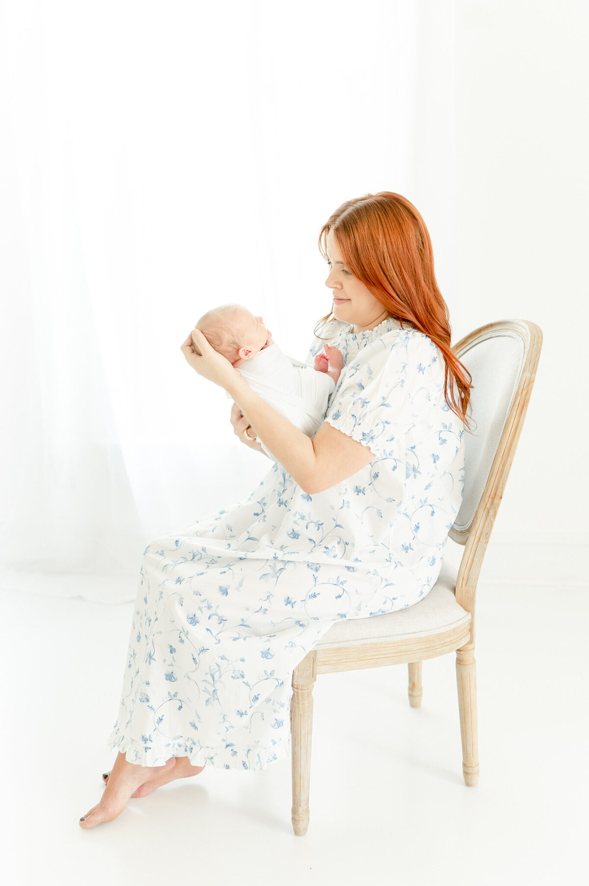 mother with red hair sits in a chair holding her newborn In Kristie Lloyd’s Nashville newborn photographer studio