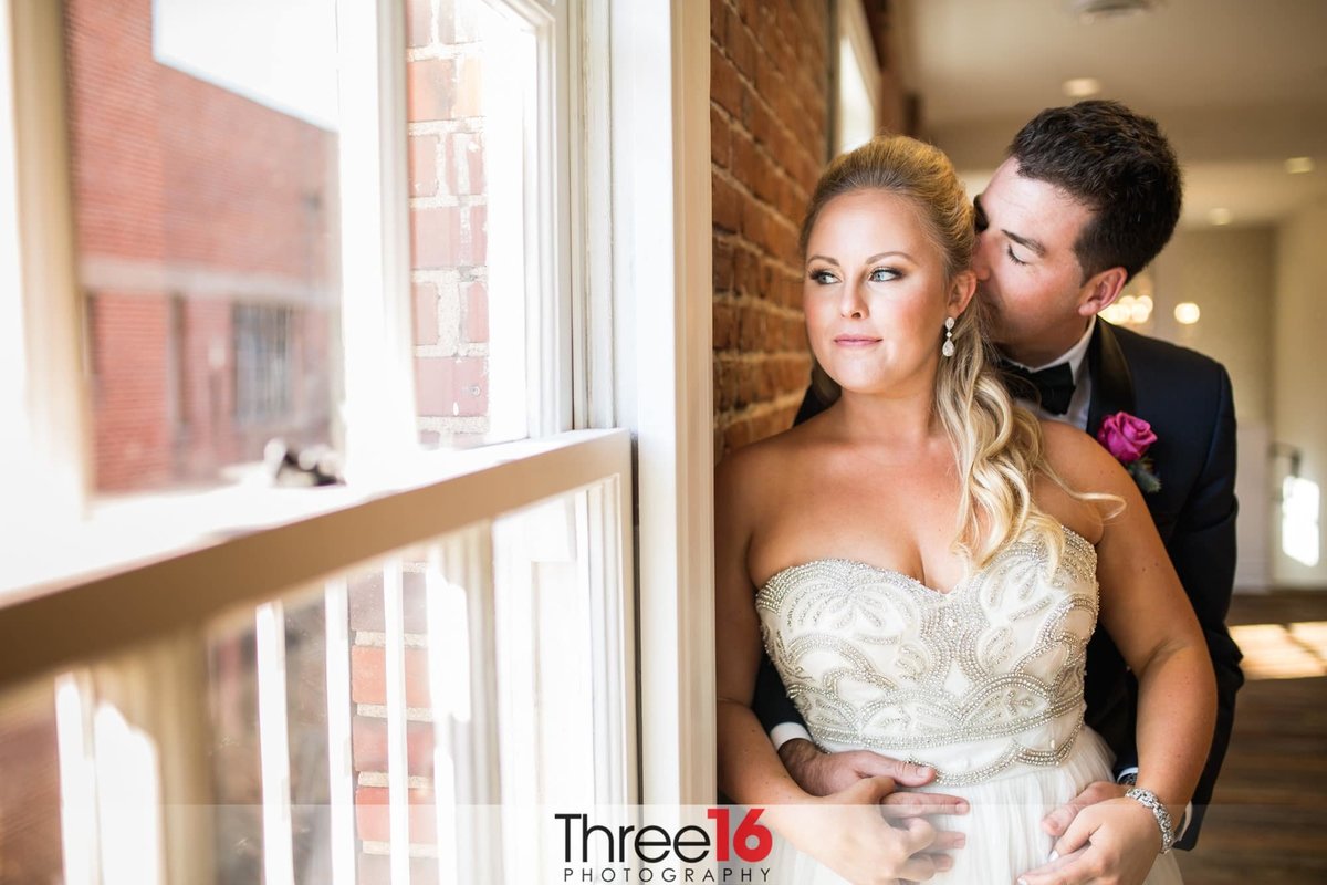 Groom holds his Bride from behind and kisses the back of her head as she looks out the window
