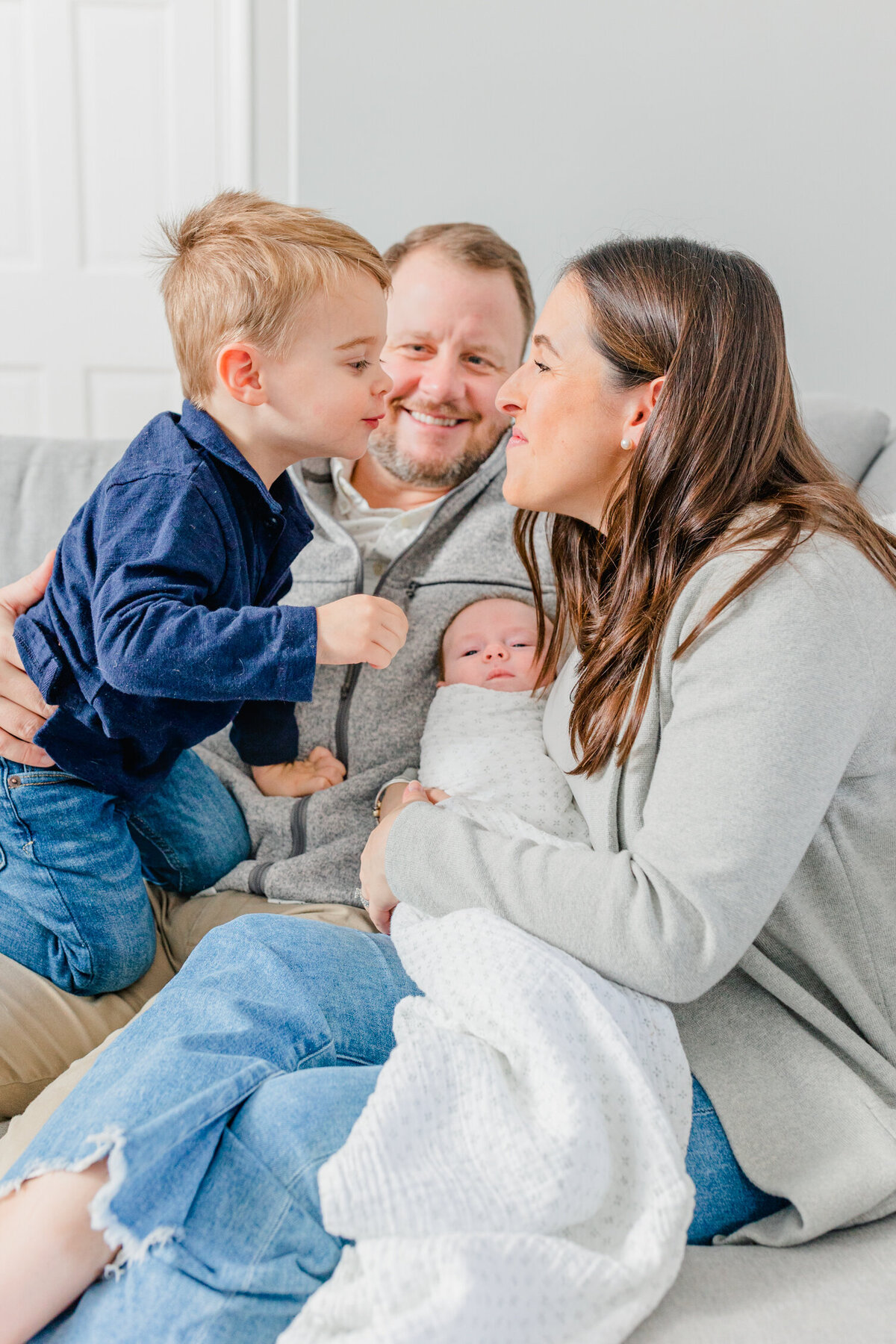 Family of four on a grey couch; dad is smiling from behind while toddler leans in to kiss his mama, who is holding a newborn baby in her lap