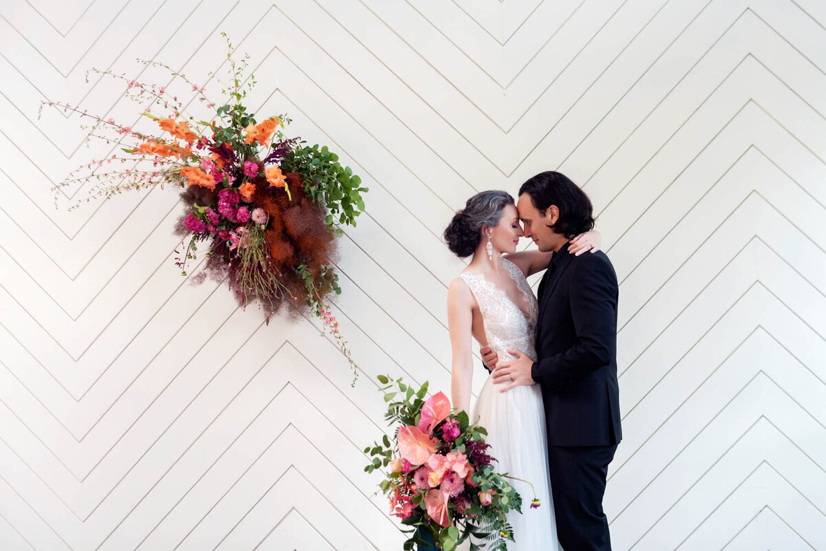 a bride and groom hug in front of a white herringbone wall with big colorful flowers