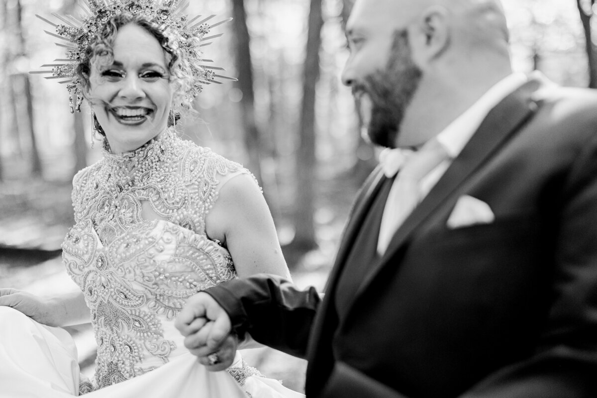 Black and white portrait of a bride and groom looking at and laughing at each other while holding hands after their wedding ceremony in DFW, Texas. The bride is on the left and is wearing a highly ornamentally decorate crown and a highly detailed and intricate dress. The groom is on the right and is wearing a dark suit with a pocket square. They are backed by a forest setting.