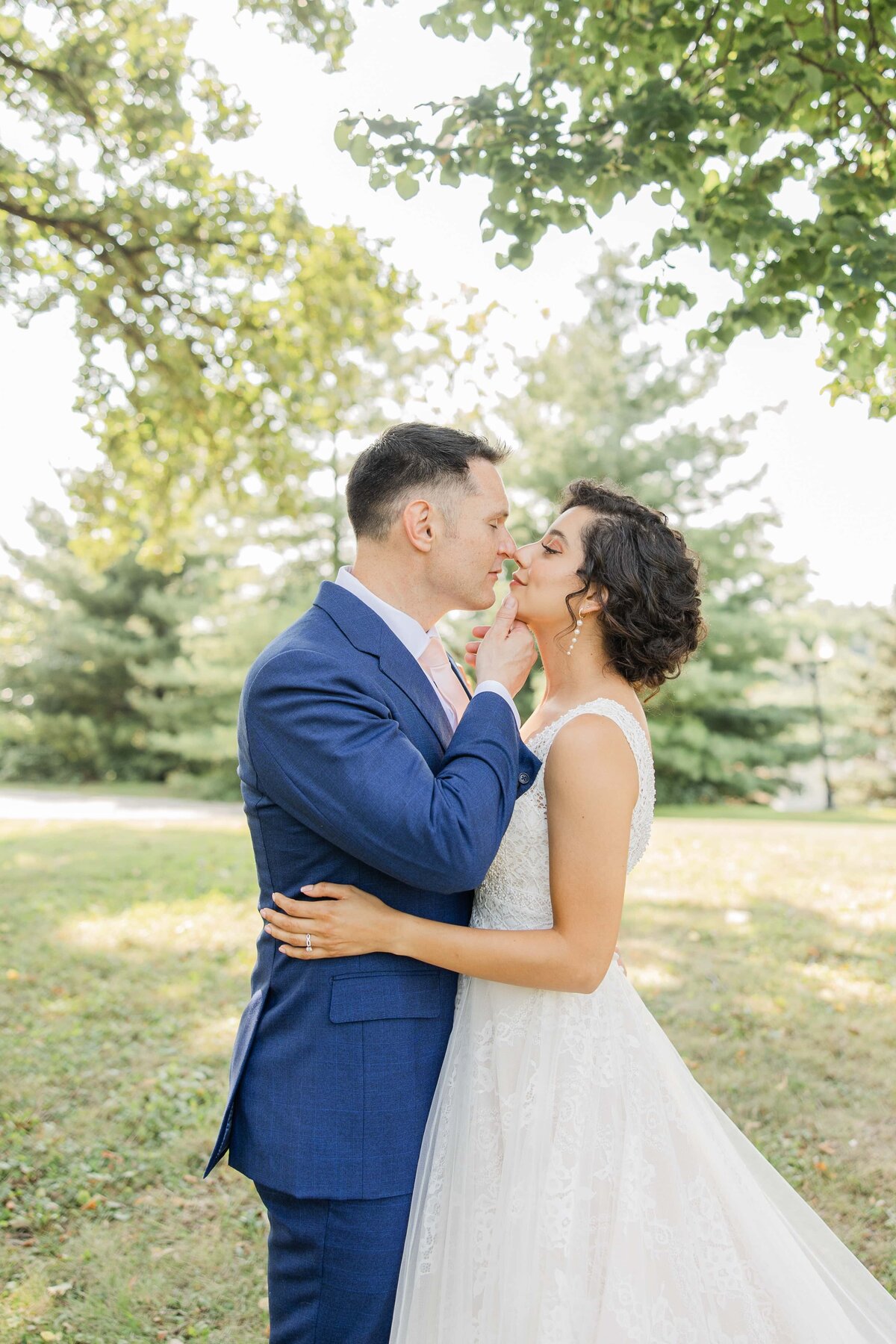 A bride in a white dress and a groom in a blue suit kissing outdoors at Park Farm Winery, surrounded by trees and sunlight.