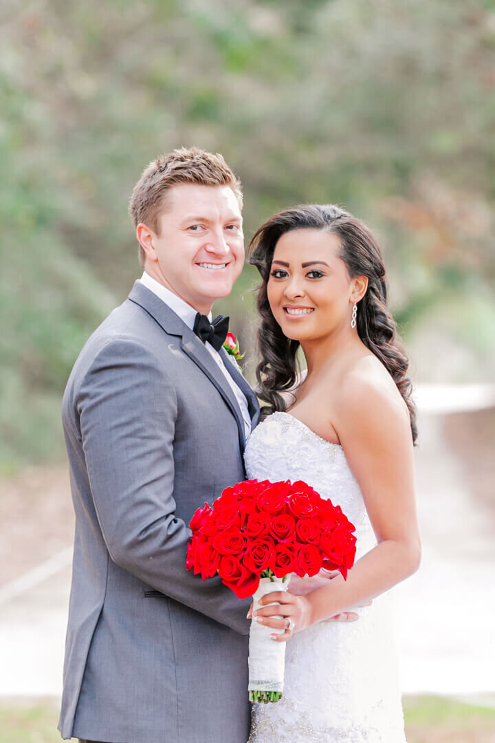Bride and Groom stand close together while she holds her bouquet of red roses in front of green trees.