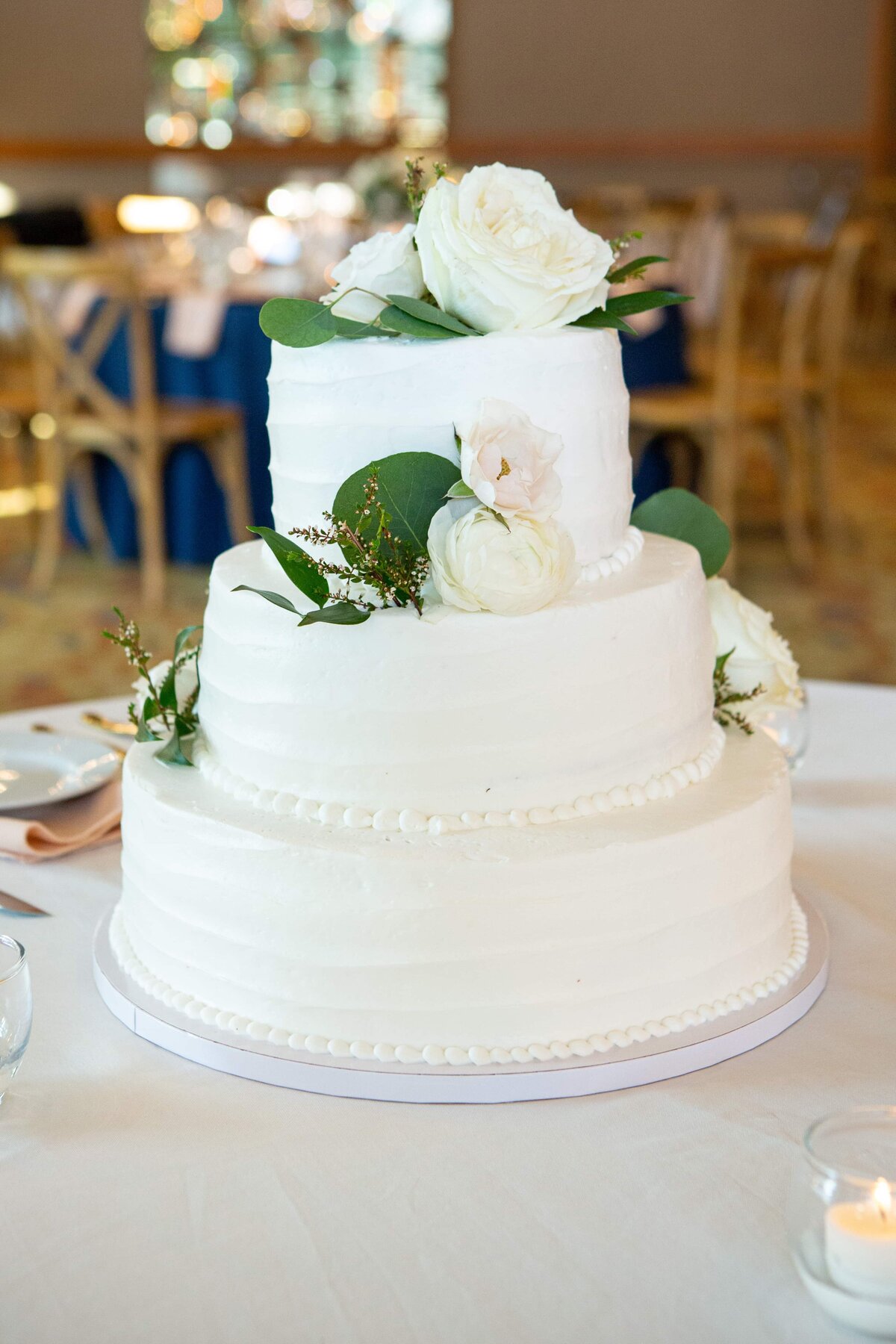A three-tiered white wedding cake decorated with fresh white roses and greenery, displayed on a table at a park farm winery wedding.