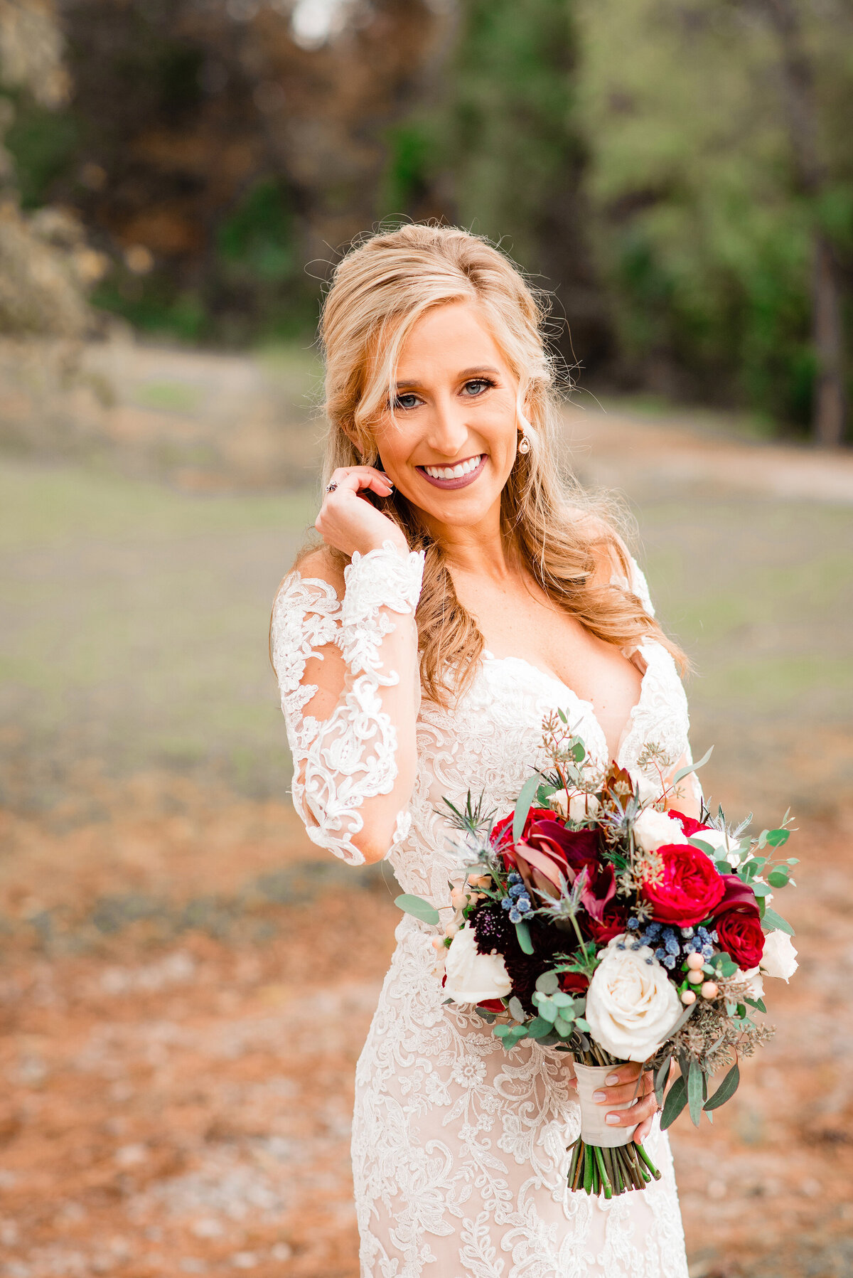 Bride adjusting hair, wearing a long sleeve lace dress and holding a cream and red bouquet