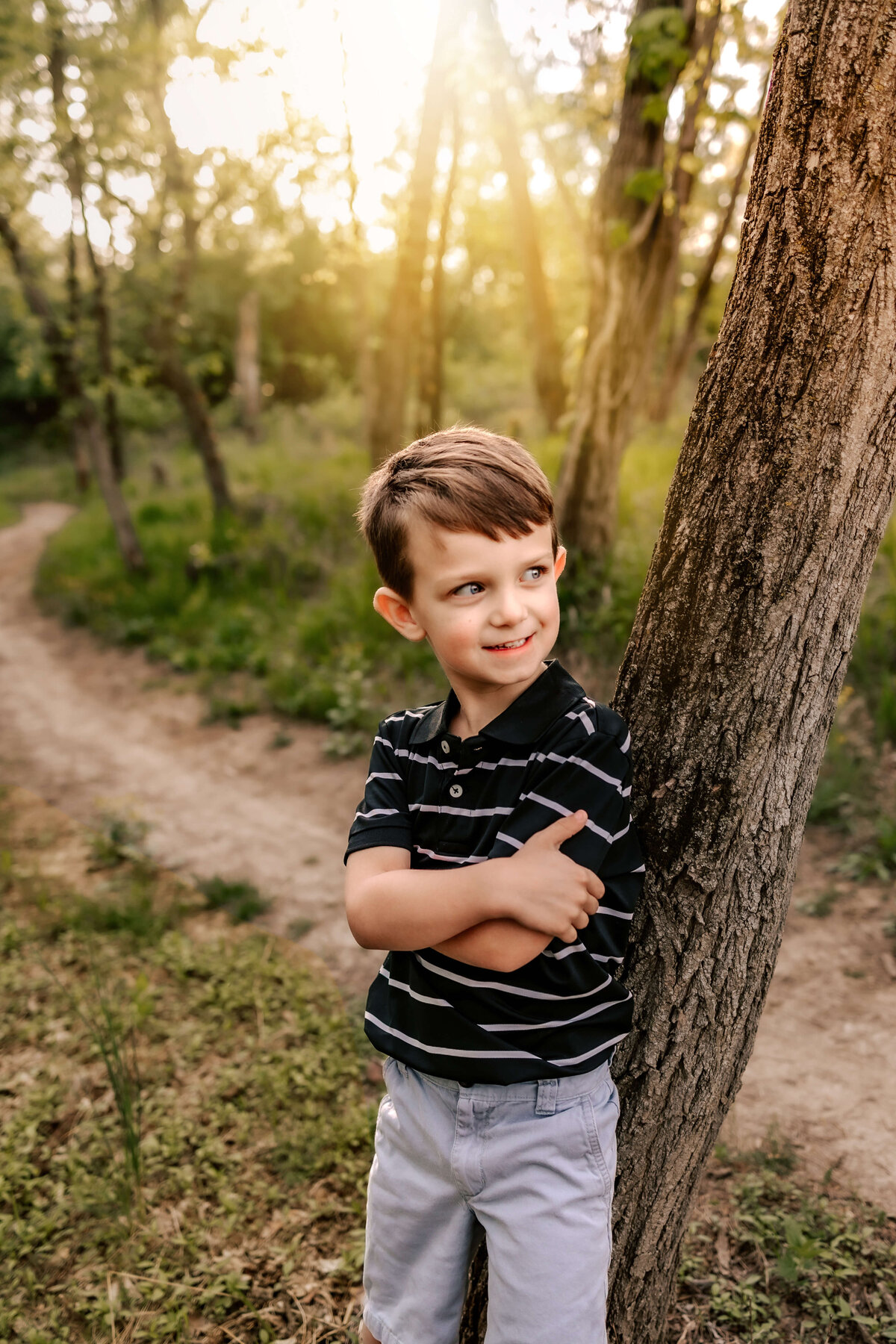 an adorable boy standing against a tree with his arms crossed and he's looking off to the distance and smiling with great joy.