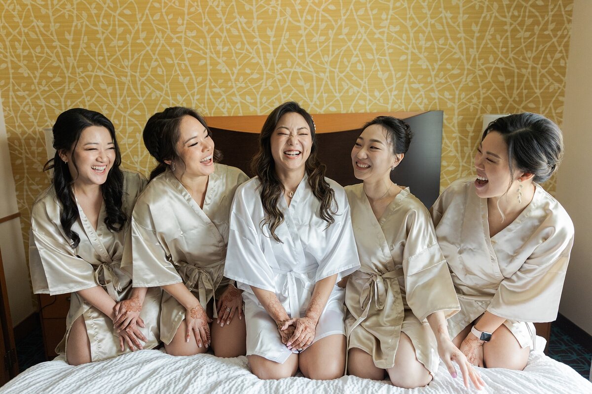 Portrait of a bride and her bridesmaids all posing together on a bed before her wedding ceremony in Dallas, Texas. The bride is in the center and is wearing a white robe. She has two bridesmaids on either side of her, and they are all wearing silvery tan robes.