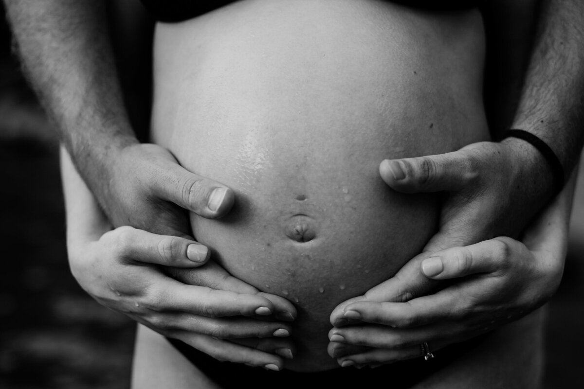 Black and white image of two sets of hands wrapped around baby belly, skin showing.