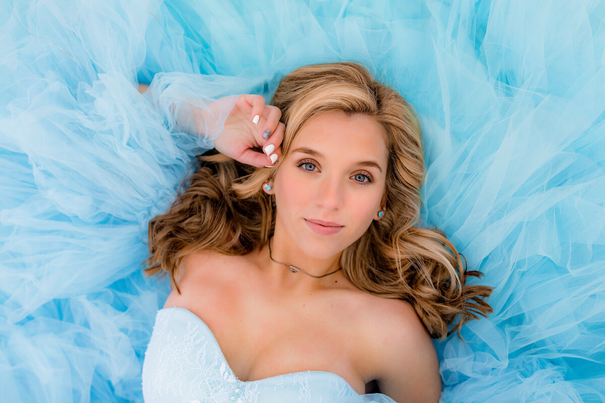 A blonde lays on the ground in a blue dress that is surrounding her face in a close up portrait.