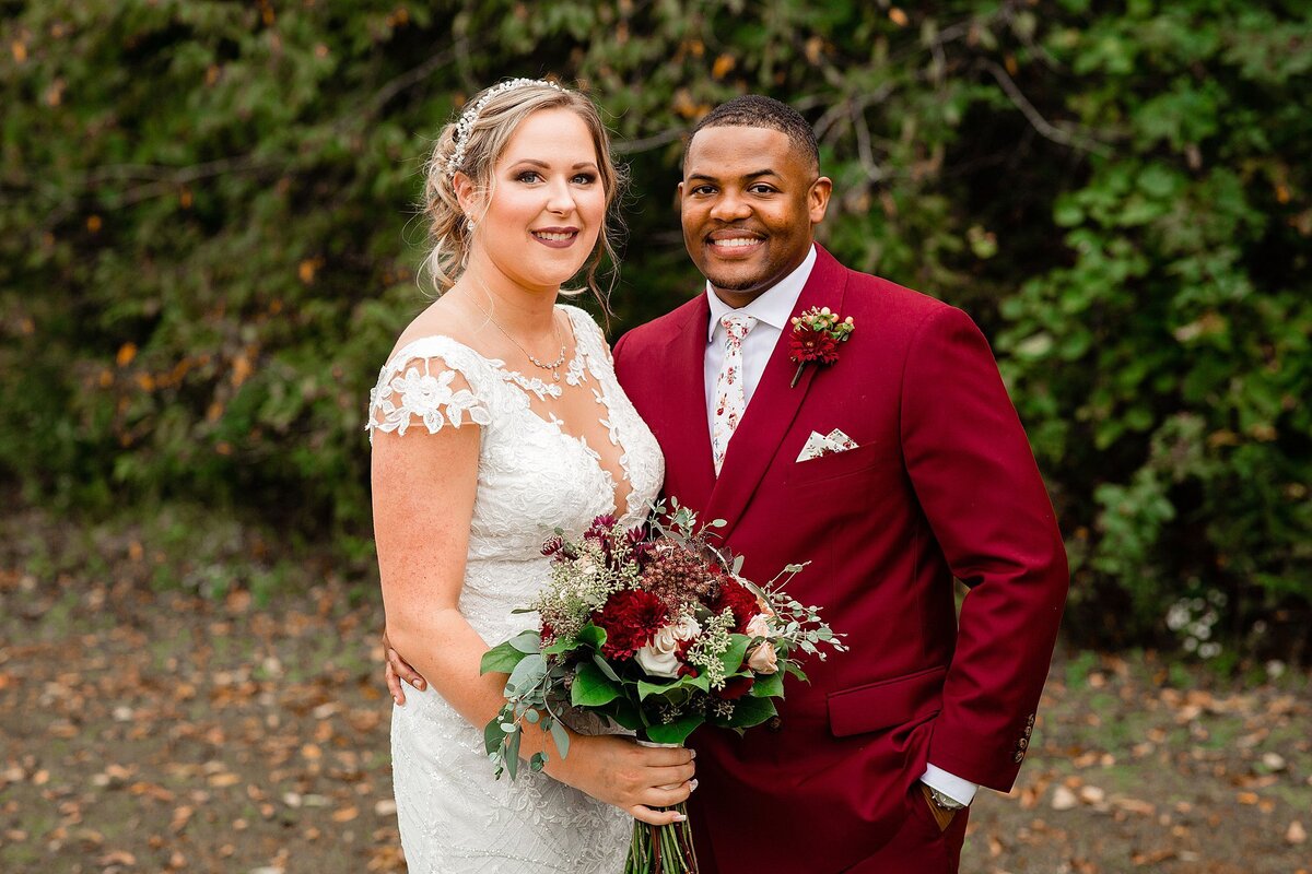 A bride wearing a short sleeves lace wedding dress with a plunging v neckline holds a bouquet of red roses, pink flowers and white roses. The African American groom is wearing a crimson red suit with a white shirt and small floral boutonniere at Wild Turkey Ranch for their Nashville wedding.