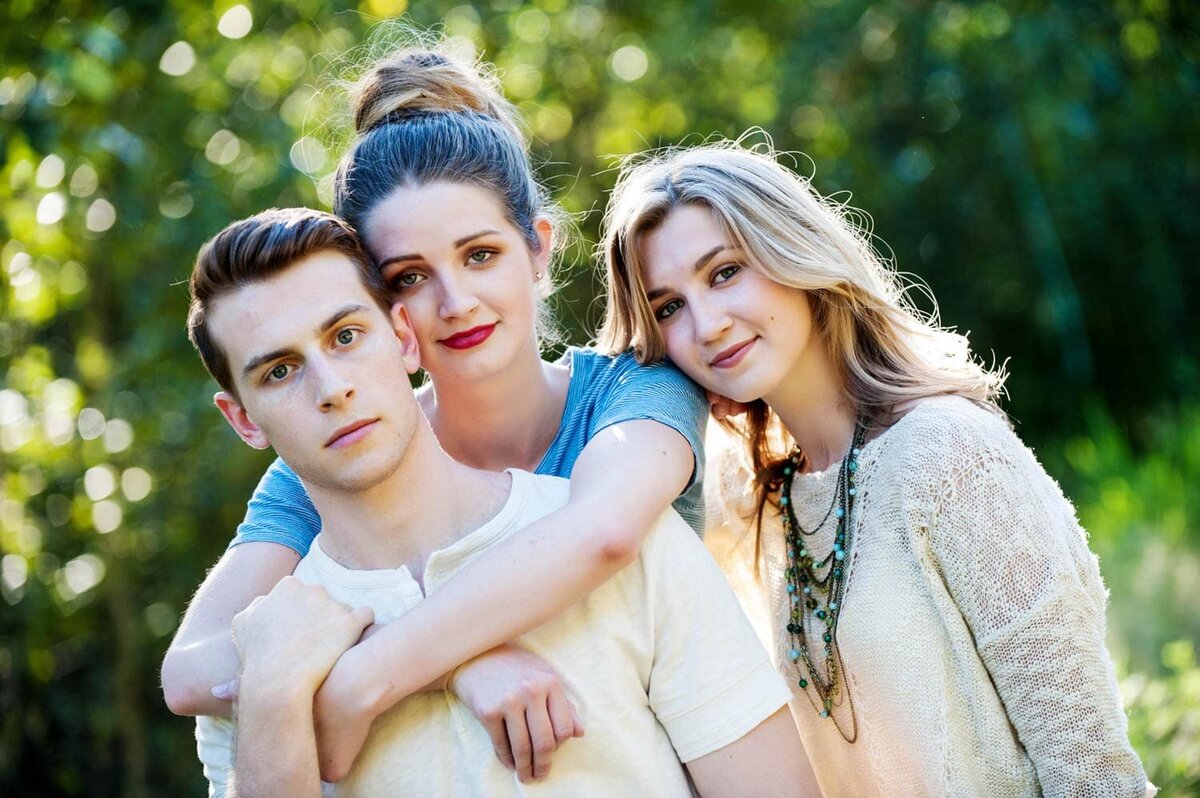 two sisters and a brother posing together in park with one sister hugging over brothers shoulders from behind.