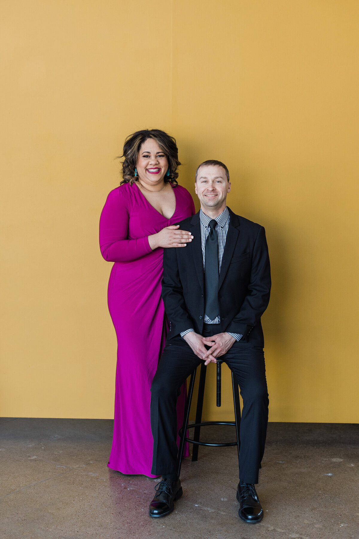 A couple posing together in front of a dark yellow background during their studio engagement session in Dallas, Texas. The woman on the right is standing and is wearing a purple dress. The man on the right is seated on a stool and is wearing a dark suit with a tie.