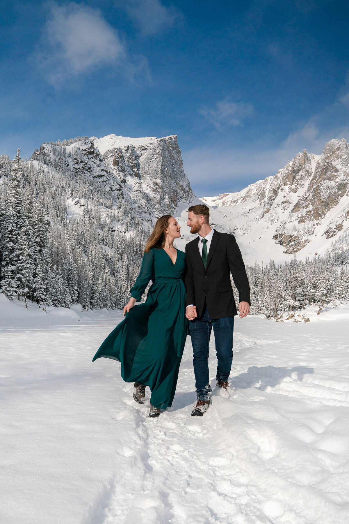 Man in suit and woman in green dress walk and smile in front of the Rocky Mountains.