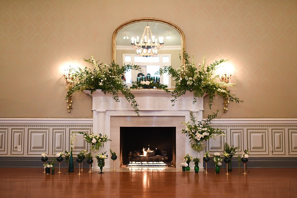 Antique mantle in Maney Hall at Oaklands Mansion decorated with large lush centerpieces of green and white flowers as a fire roars in the fireplace.