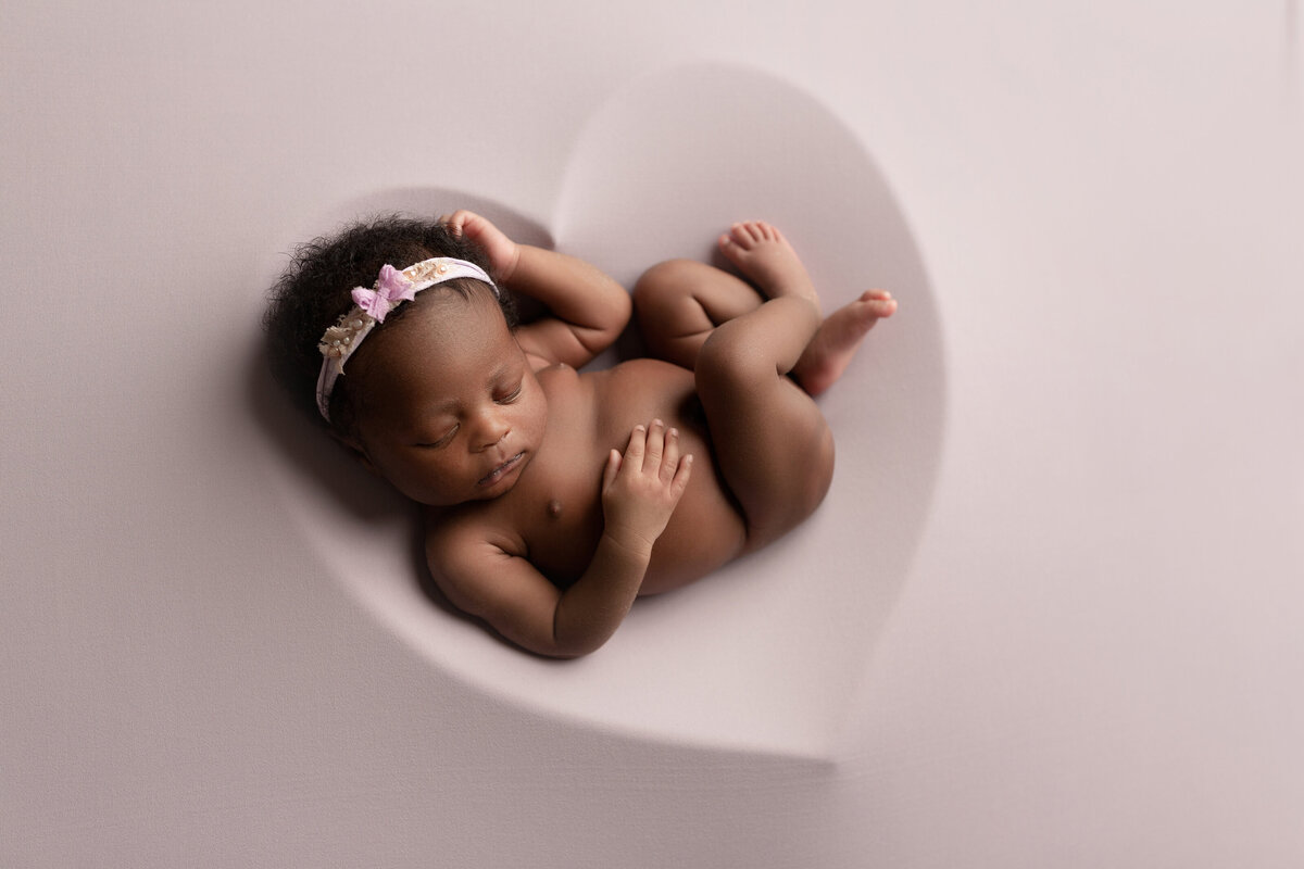 Black baby girl resting atop of a blush stretch fabric with the impression of a heart beneath her. Baby is bare, resting one hand under her head and the other on her belly. Legs are folded. Baby is wearing a soft pink headband.