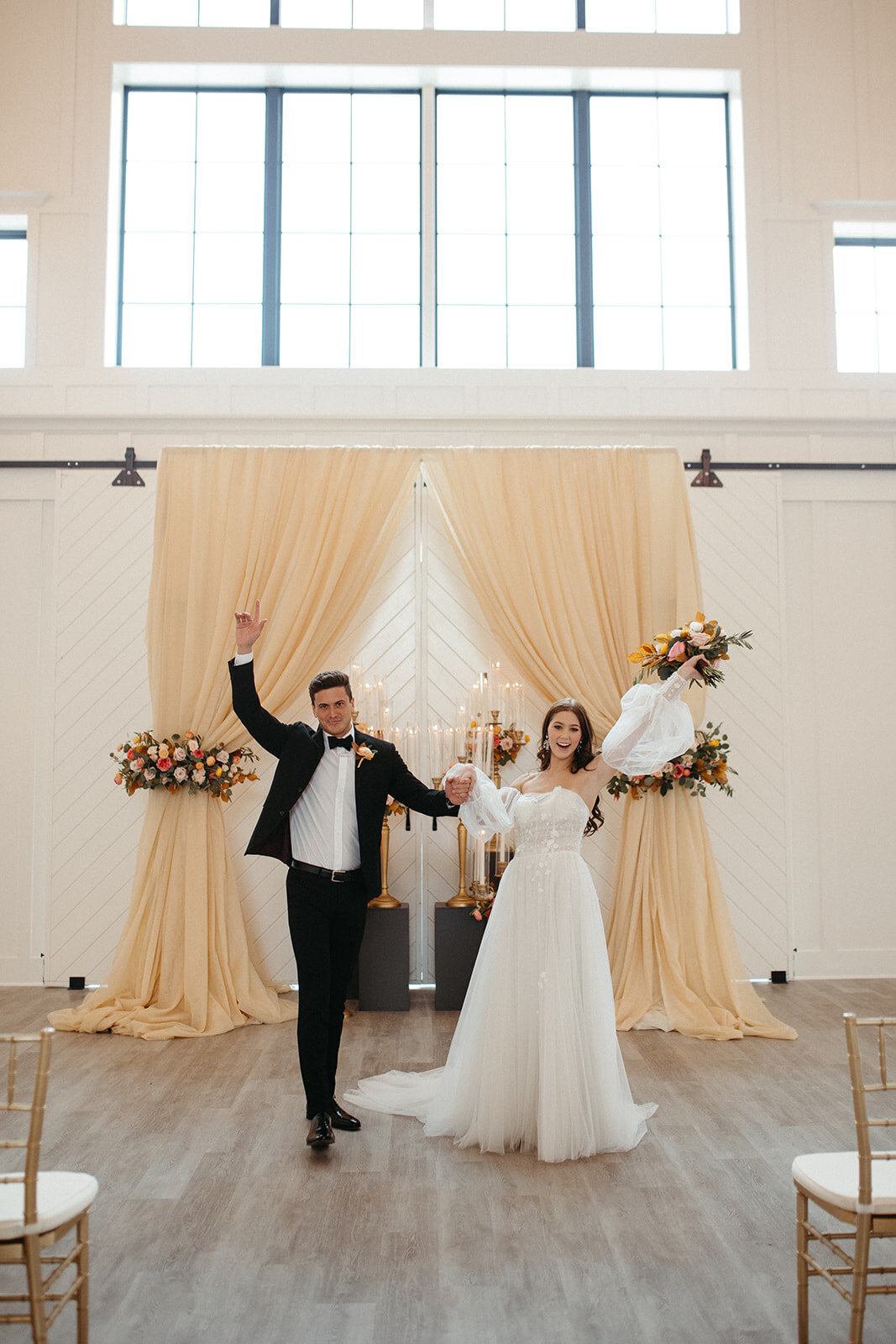 A bride and groom wearing a white wedding gown and black tuxedo pose with one hand in the air in front of peach curtains.