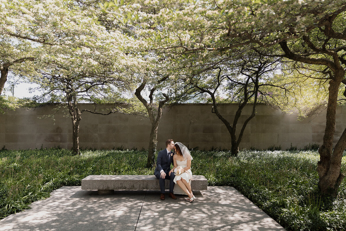 Just Married photo session couple sits on bend in the Chicago Art Institute garden with beautiful trees and light. Groom is kissing the brides forehead.