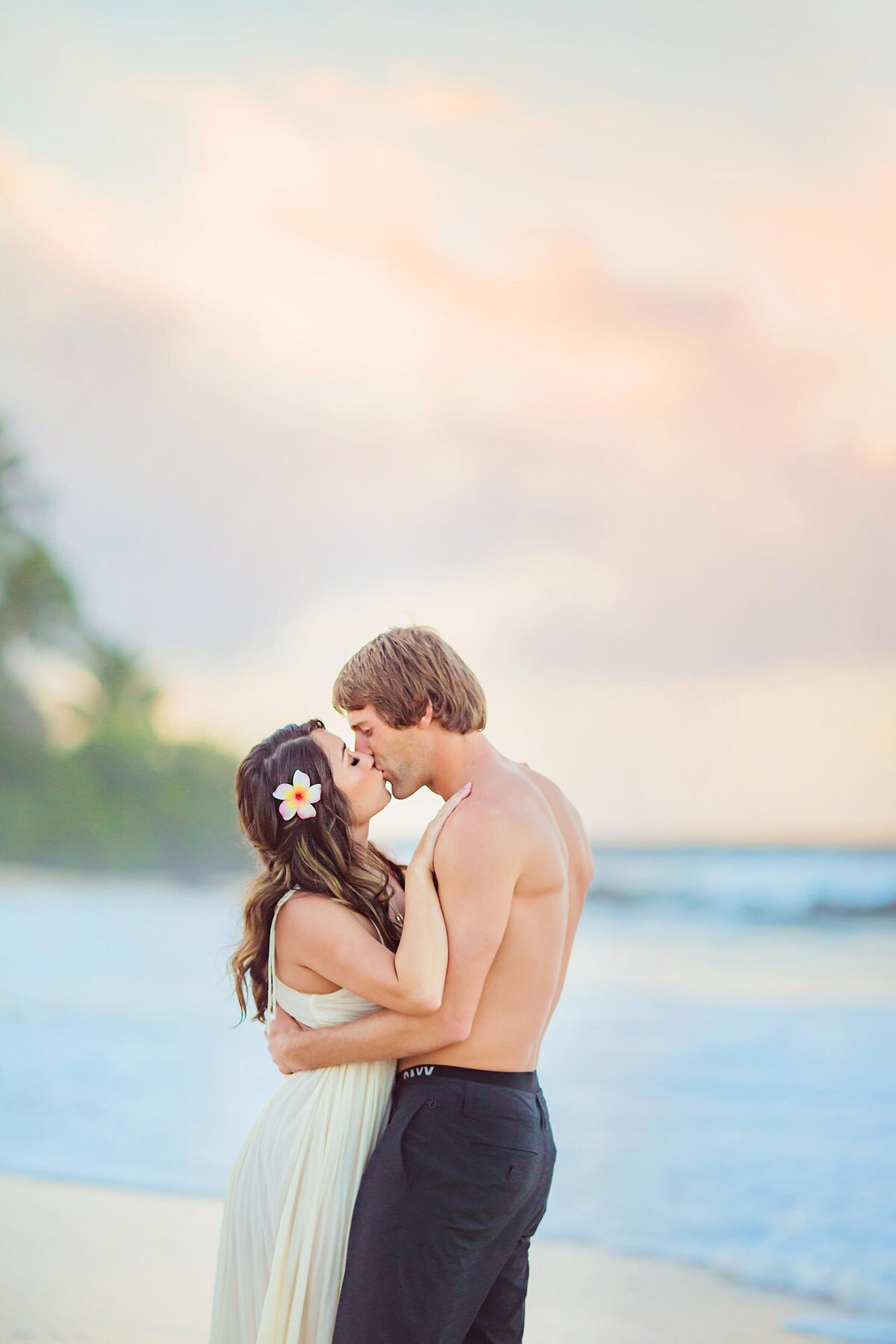 Woman wearing a cream dress wearing a flower in her hair is kissed by her shirtless boyfriend and they are photographed by Love + Water Photography
